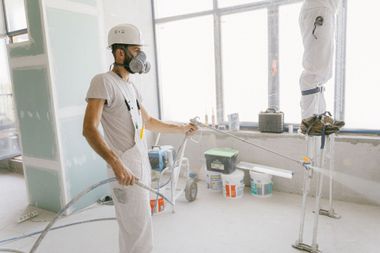 Worker in a white protective suit and respirator stands in a bright room during painting or renovation.