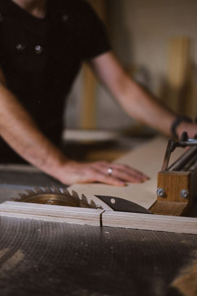 Person working at a table saw in a woodworking shop, hands guiding wood past the blade.