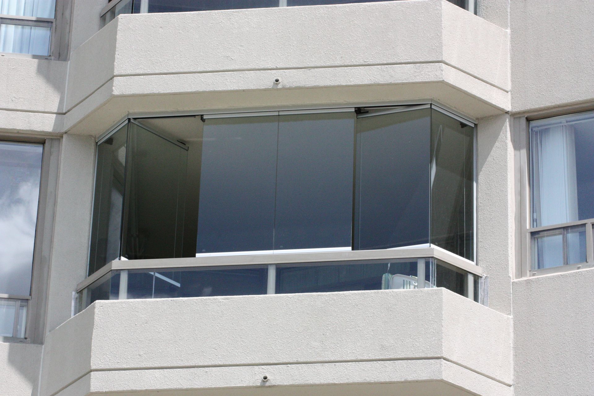 Glass-enclosed balcony on a concrete building.  Gray-tinted glass windows and white concrete structure.