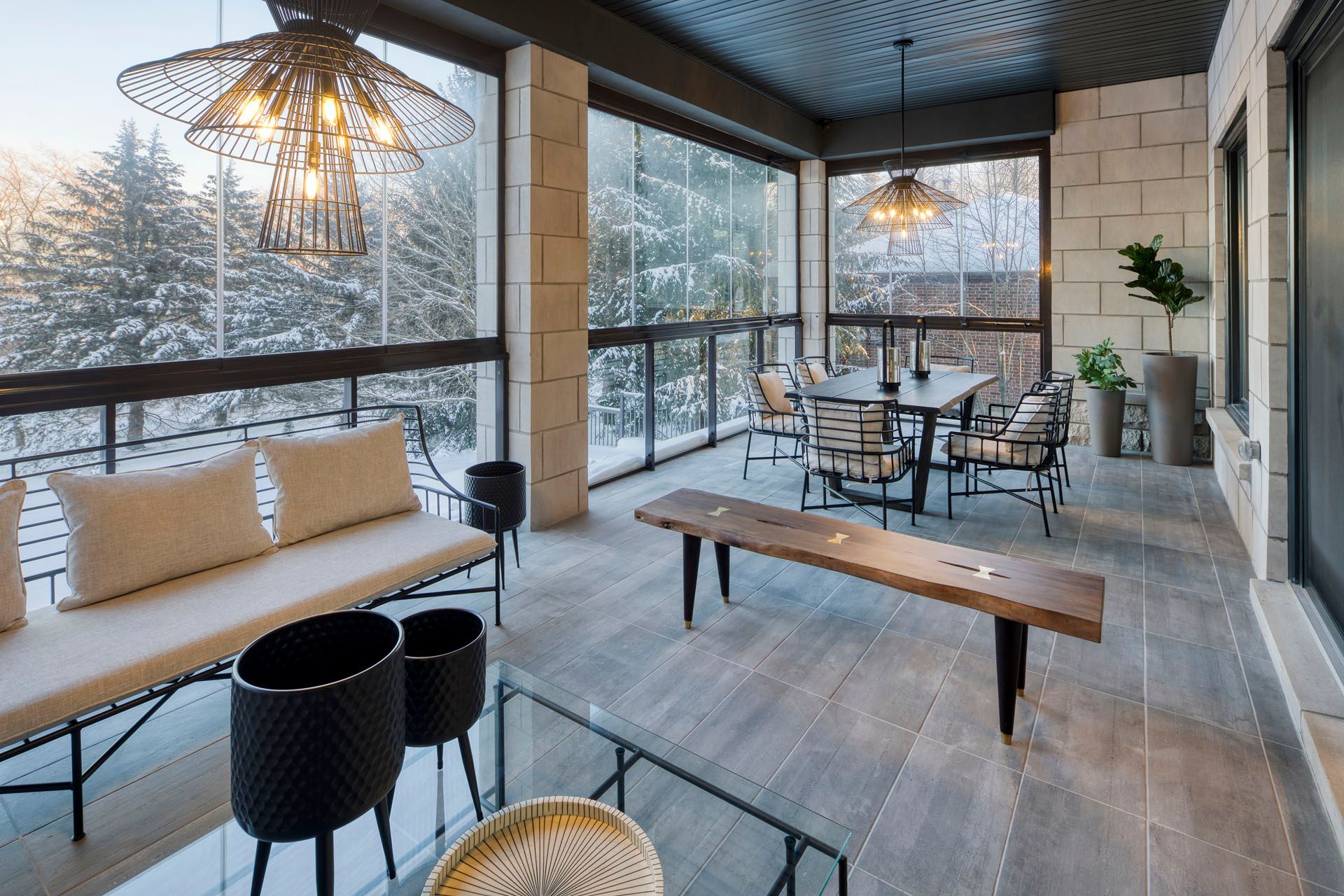 Enclosed porch with snow-covered trees visible through glass walls, furnished with seating, table, and decorative lighting.