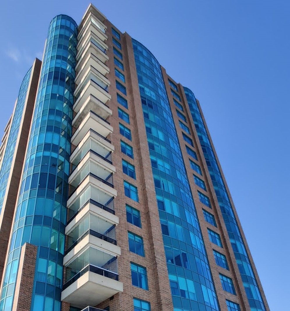 Tall building with blue glass facade, brown brick sides, white balconies, against a clear blue sky.