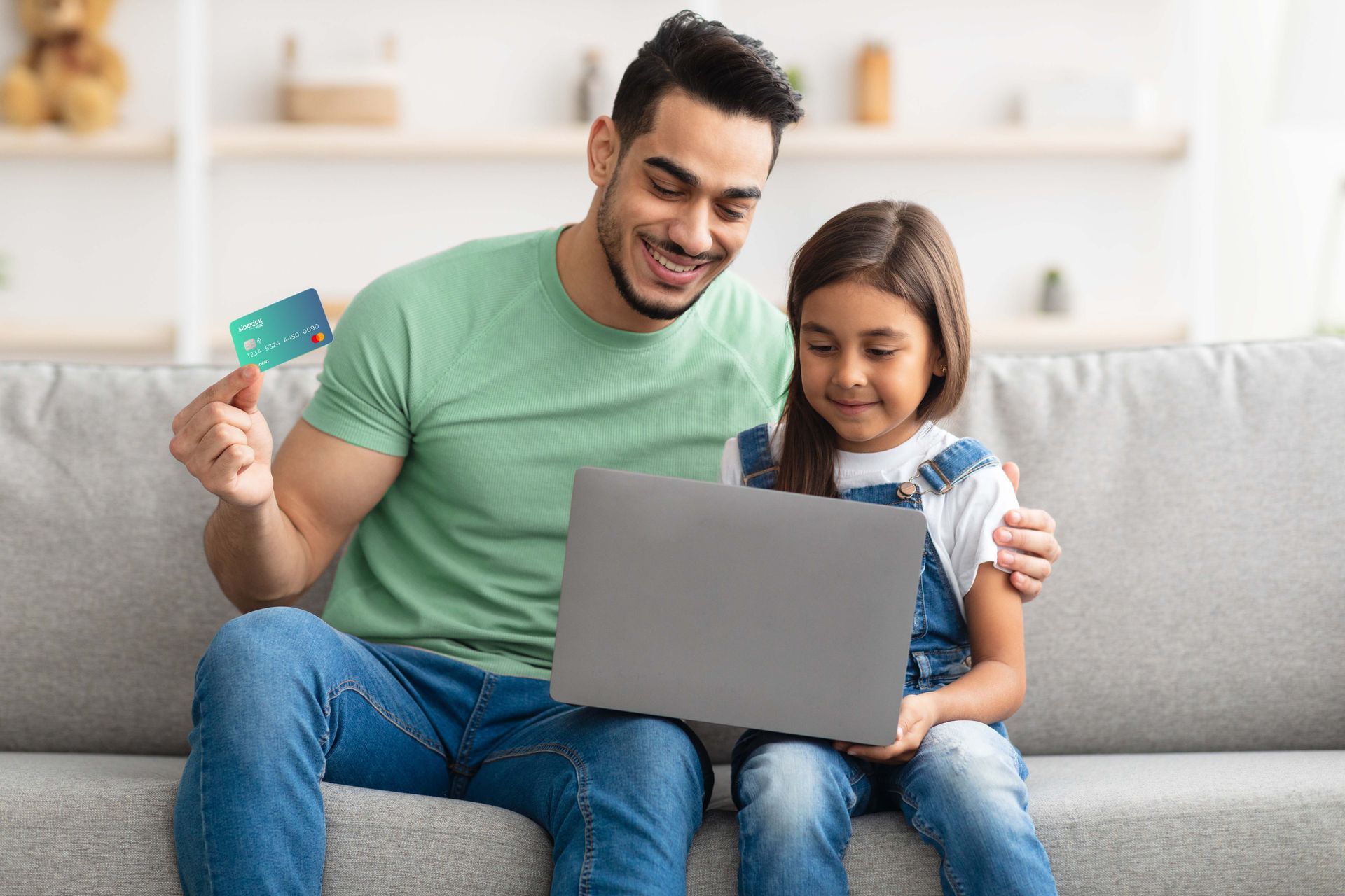 Smiling man and child using a laptop on a couch, holding a credit card in a bright living room