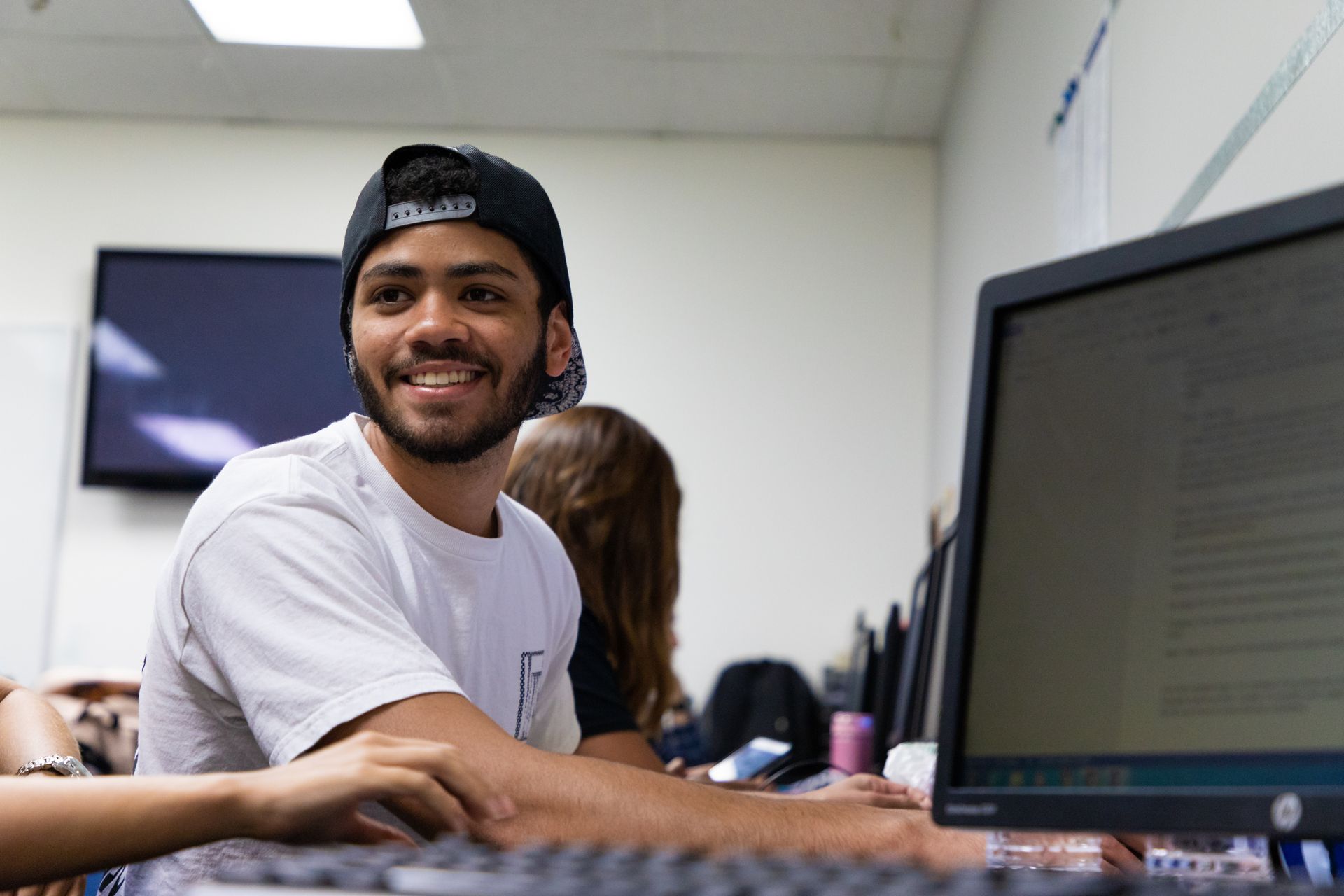 Smiling man typing at a computer in an office, wearing a black cap and white shirt.