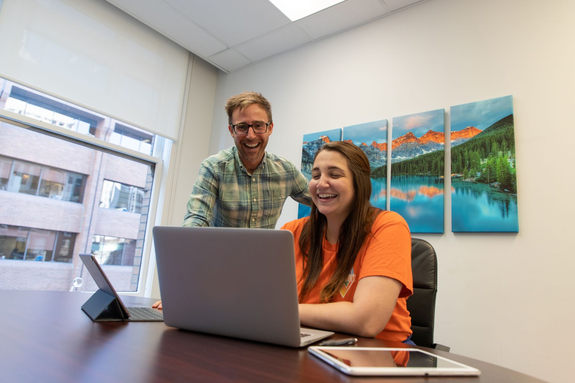 Two coworkers smiling at a laptop in a bright office with windows and wall art.
