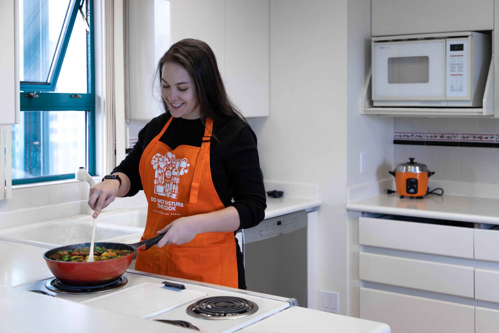 Person cooking at a stovetop in a bright kitchen, wearing an orange apron and stirring a pan.