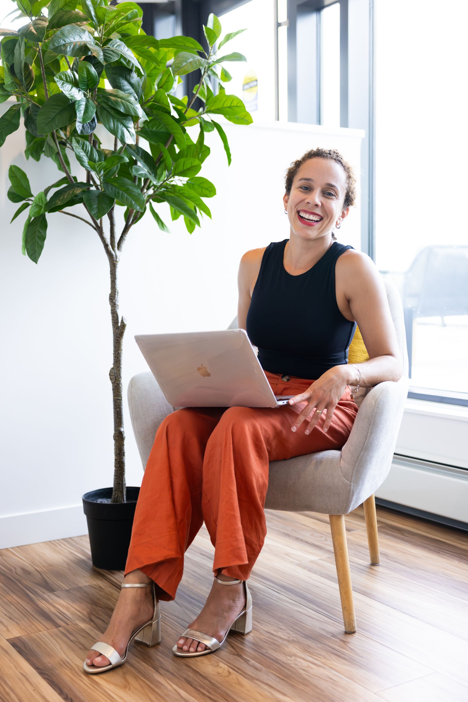 Woman seated with a laptop beside a potted tree in a bright room, smiling near a window