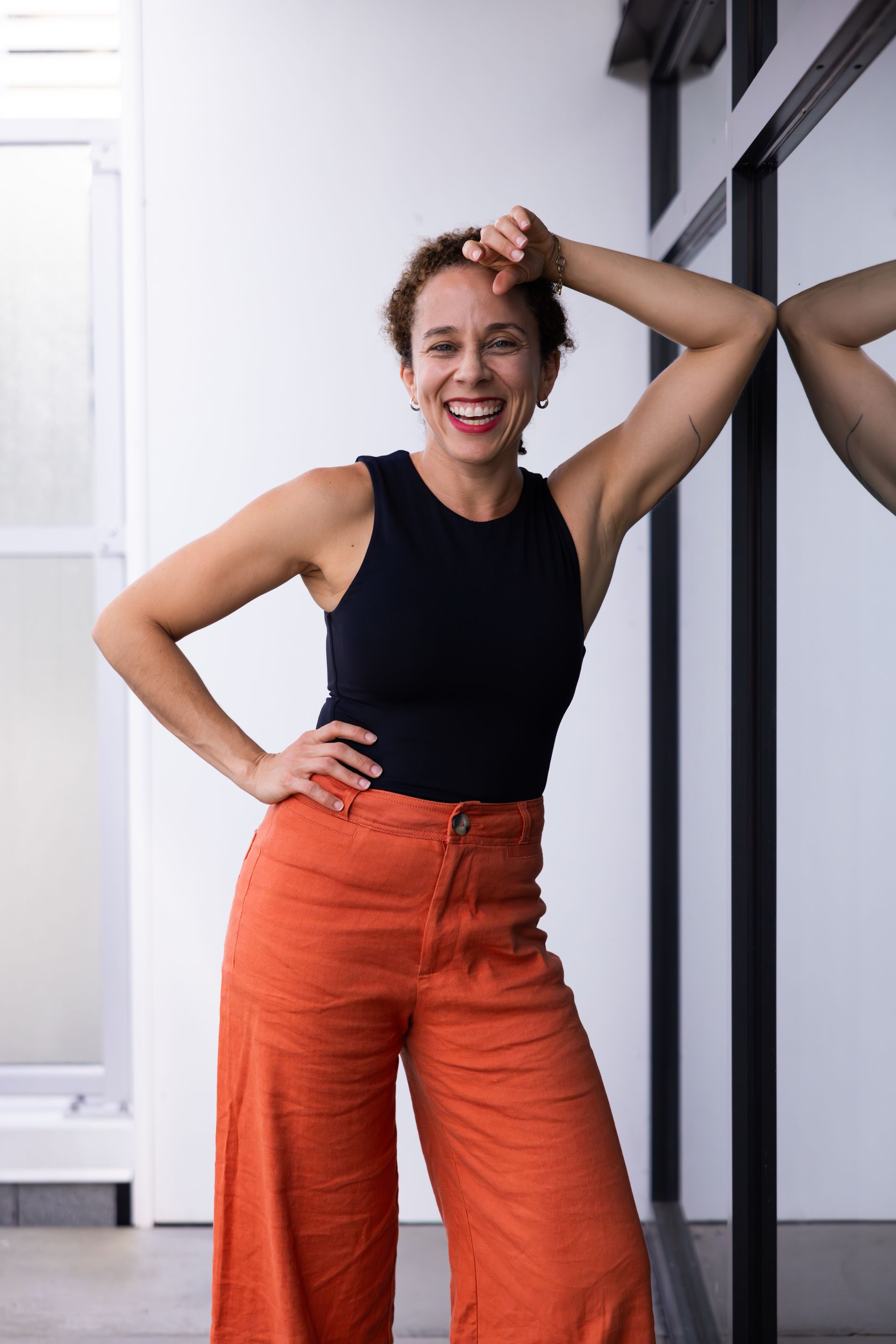 Smiling woman in black top and orange pants posing indoors by a mirror