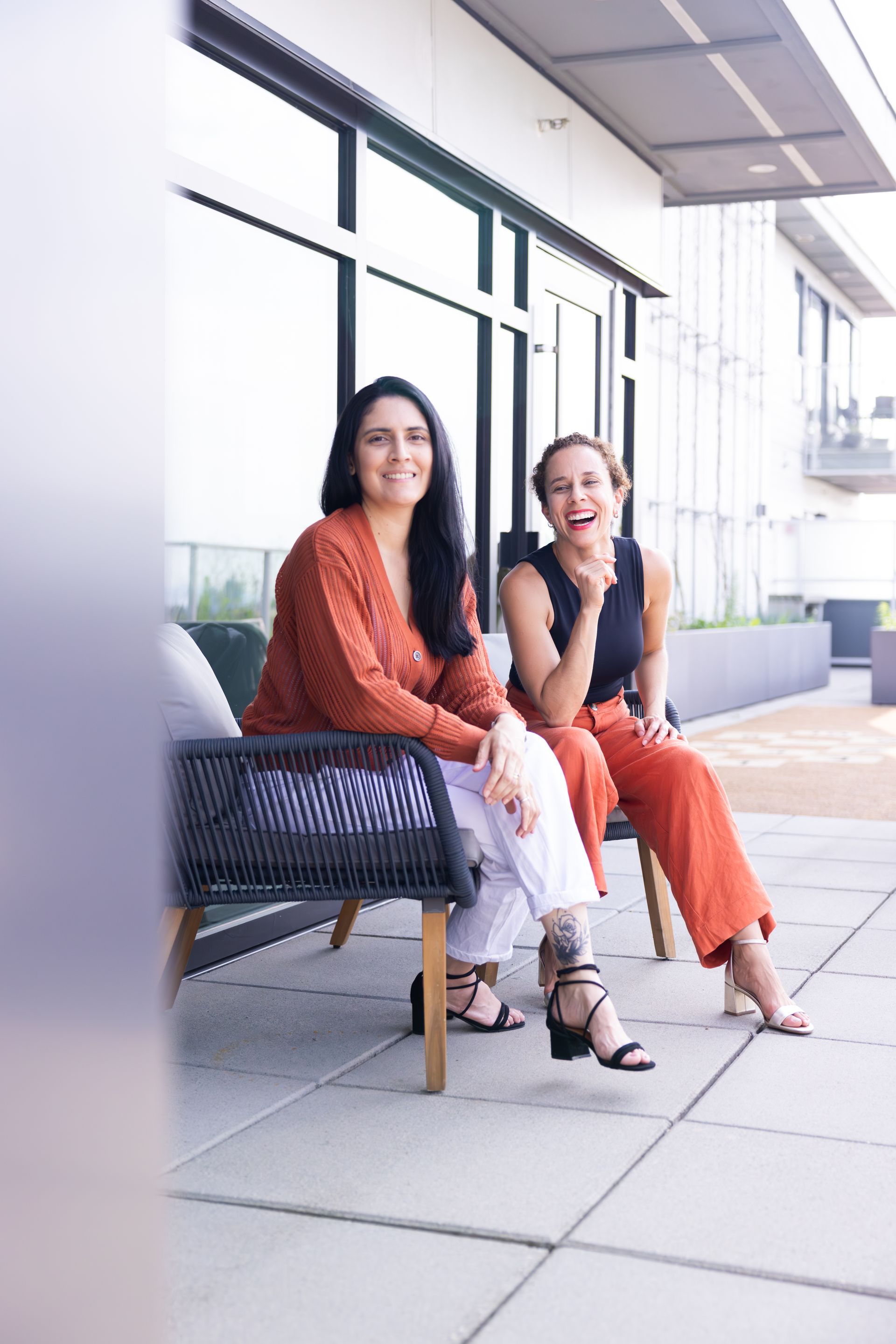 Two women sitting on a sidewalk outside a building, smiling and chatting, one in orange and one in white with a black handbag