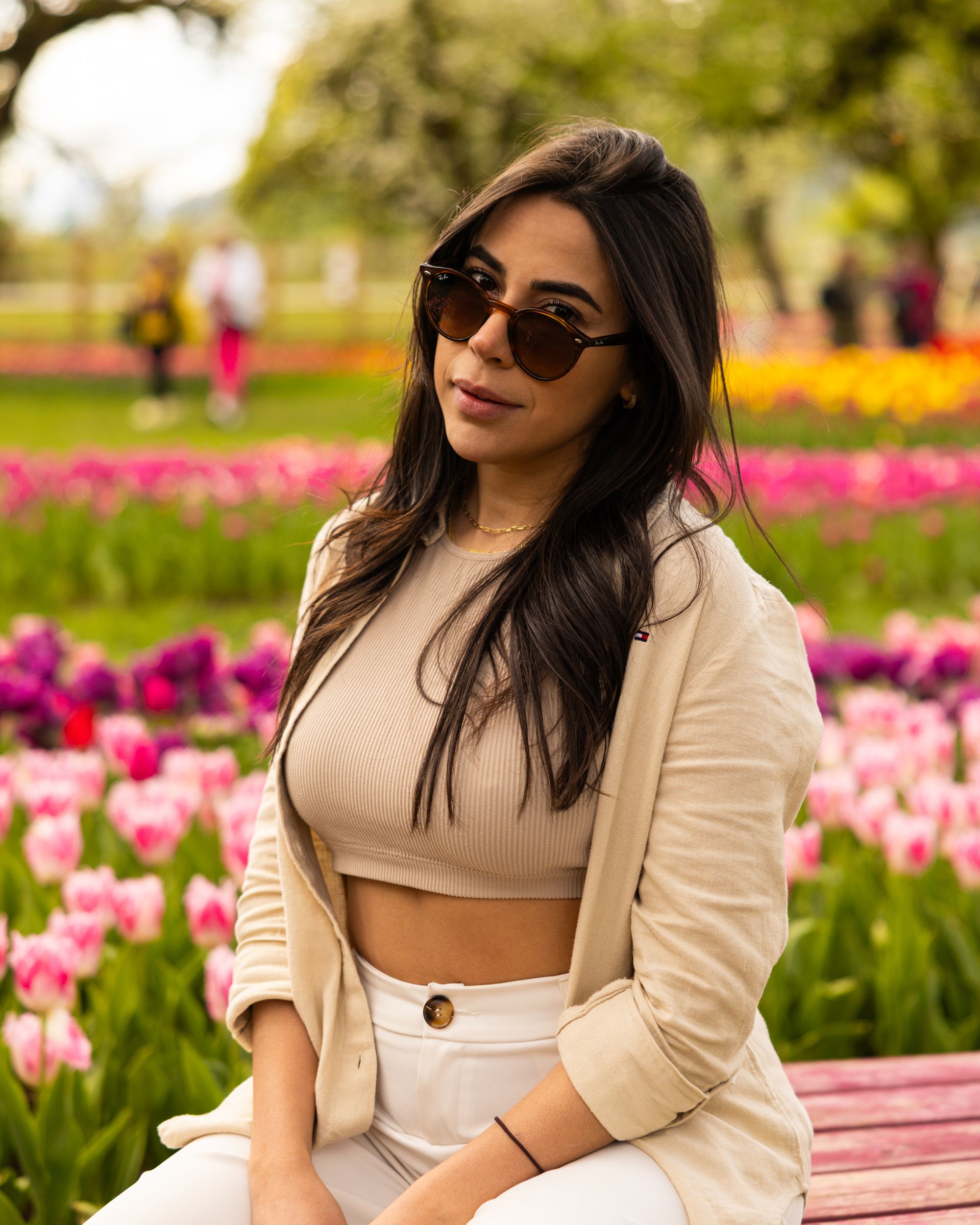 Woman in sunglasses standing by a pink flower bed in a garden, wearing a beige top and white pants.