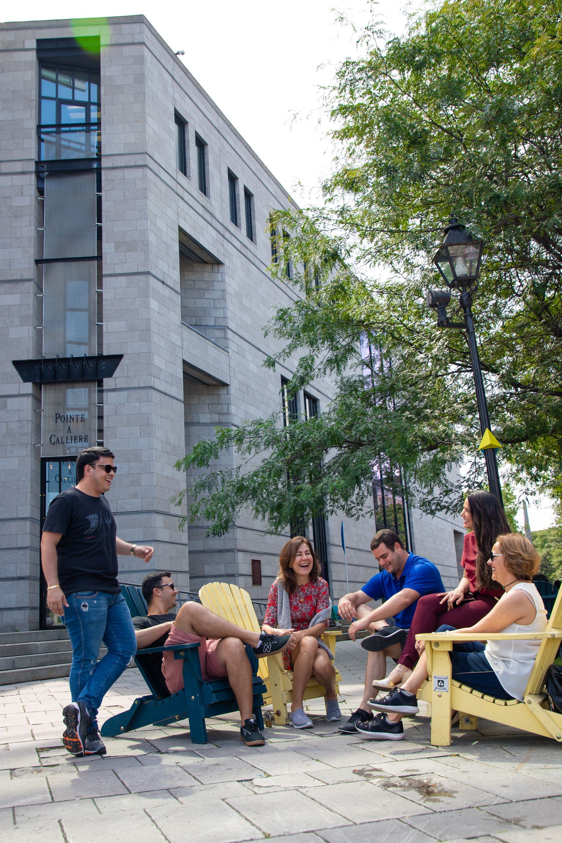 People sitting and chatting on benches outdoors beside a gray building and a tree.