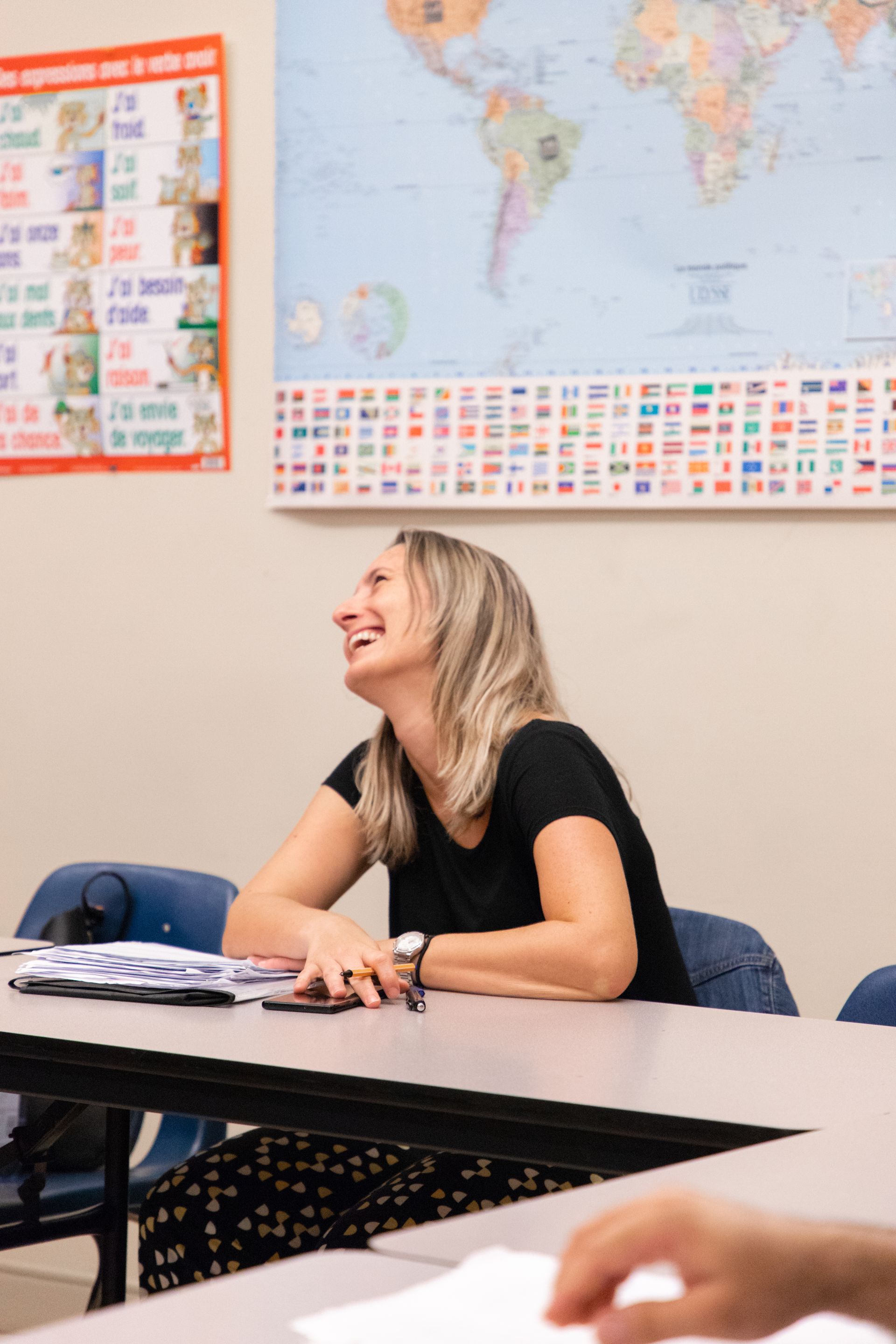 Woman smiling at a classroom desk with world maps and posters on the wall