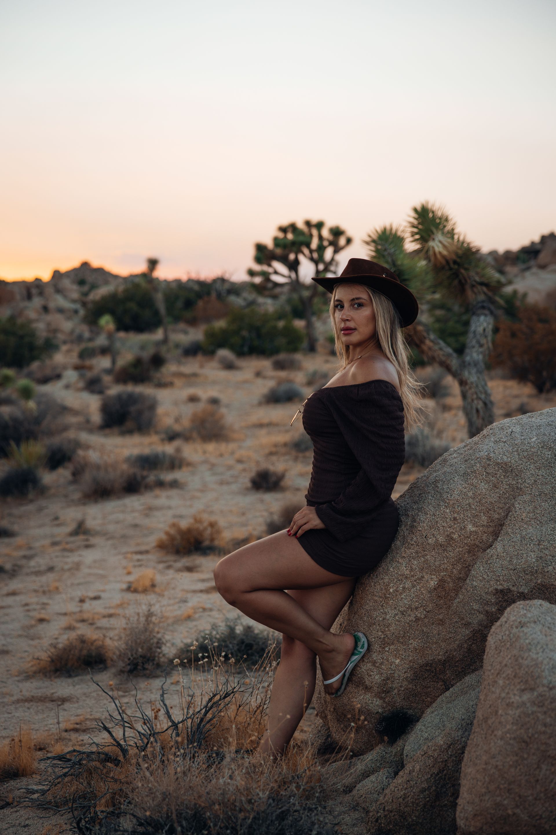 Woman in a black dress and hat posing on a rock in a desert at sunset.
