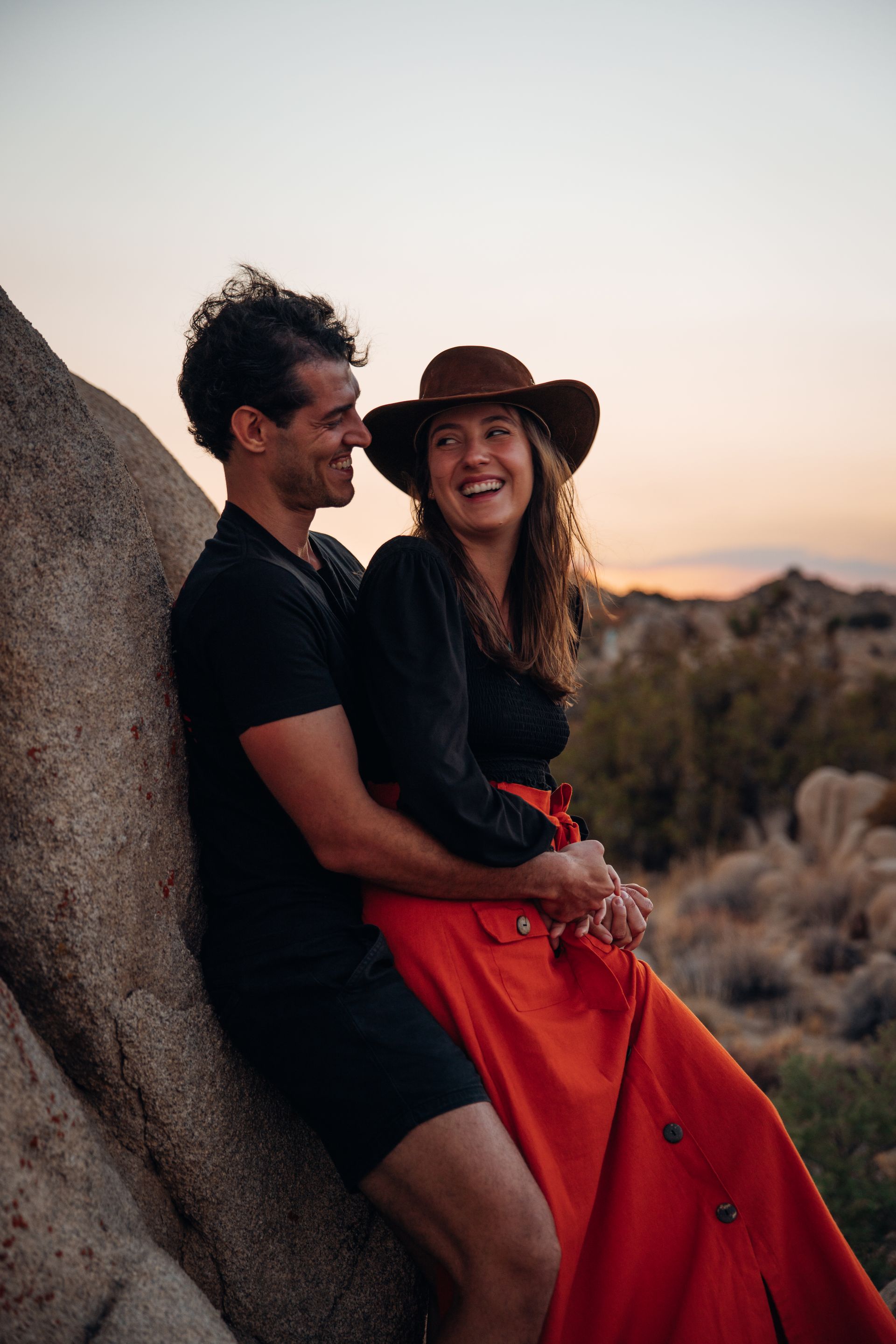 Couple sitting on a rock at sunset, smiling; woman in red dress and hat, man in black shirt.