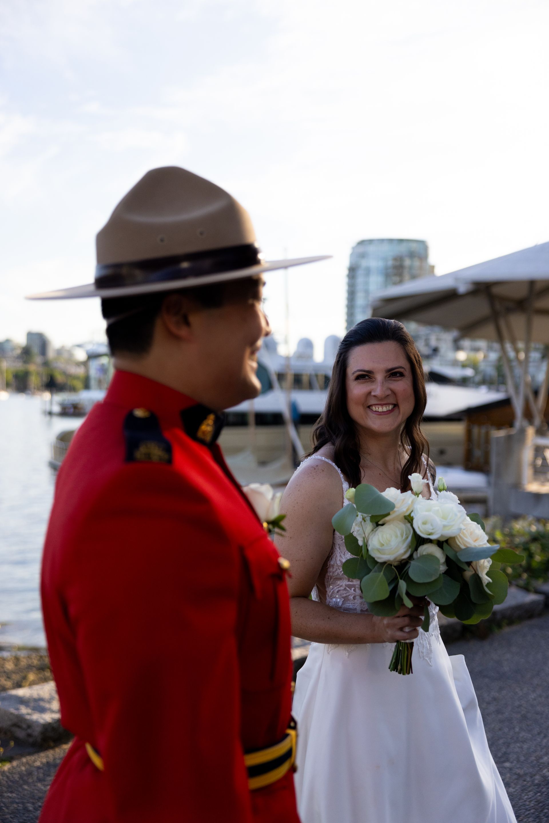 Bride smiles while holding bouquet beside a mounted officer by the waterfront