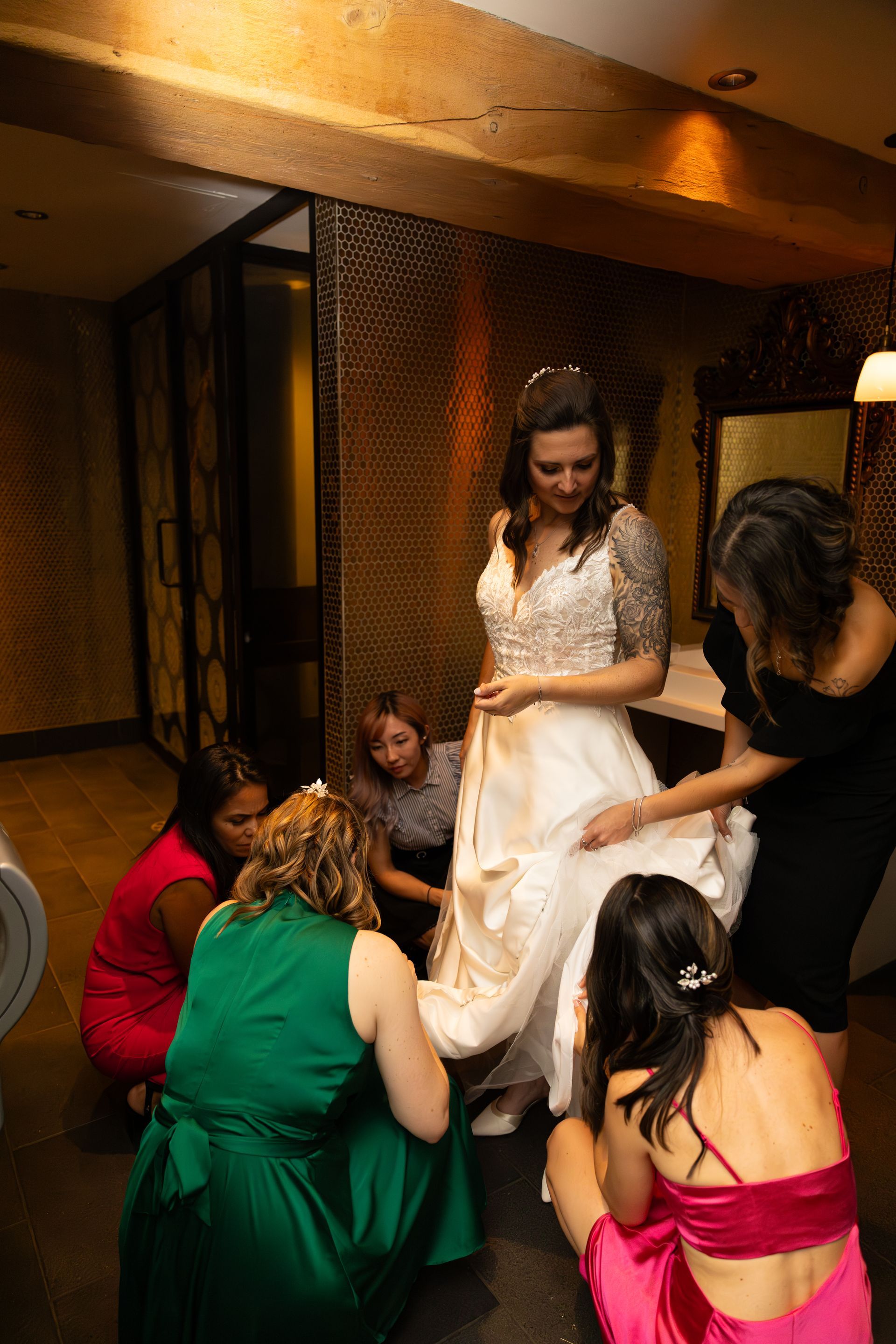 Bride in white dress stands with bridesmaids helping adjust her gown in a warmly lit room.