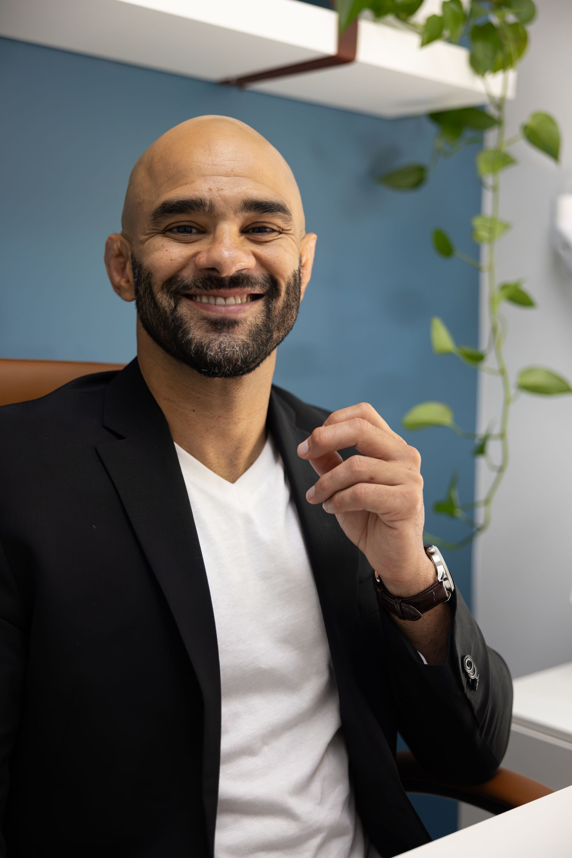 Smiling man in a black blazer and white shirt sitting indoors near a plant, holding a small object.