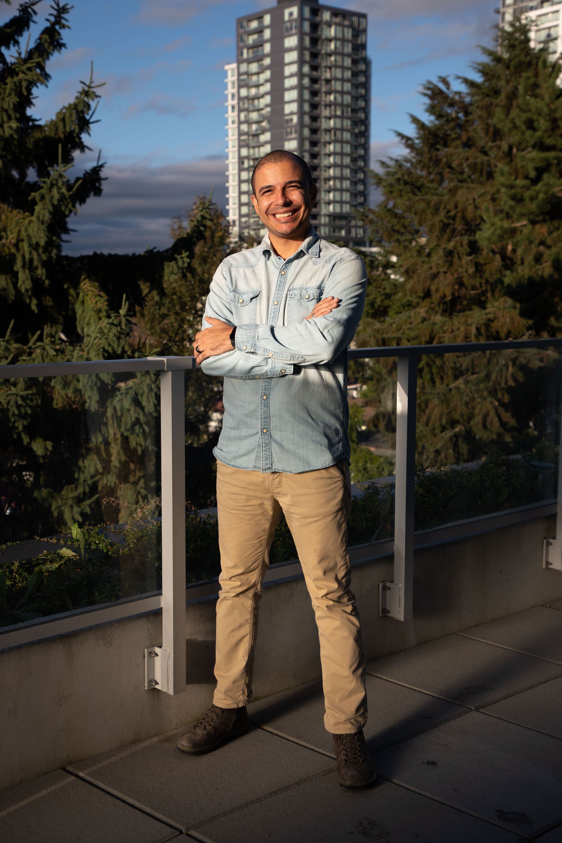 Man standing on a balcony with arms crossed, wearing a light shirt and tan pants, with a tall building and trees behind him.
