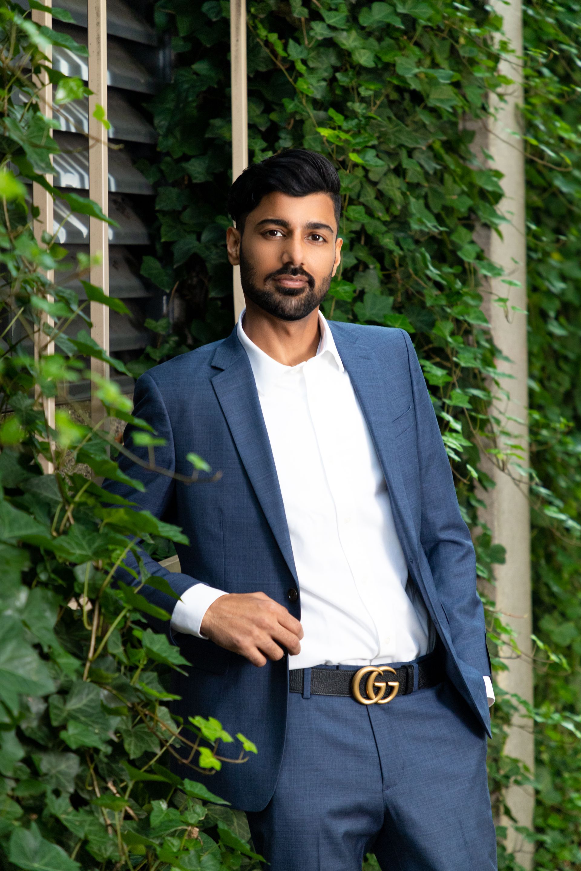 Man in a blue suit leaning by a leafy wall, wearing a white shirt and gold belt buckle