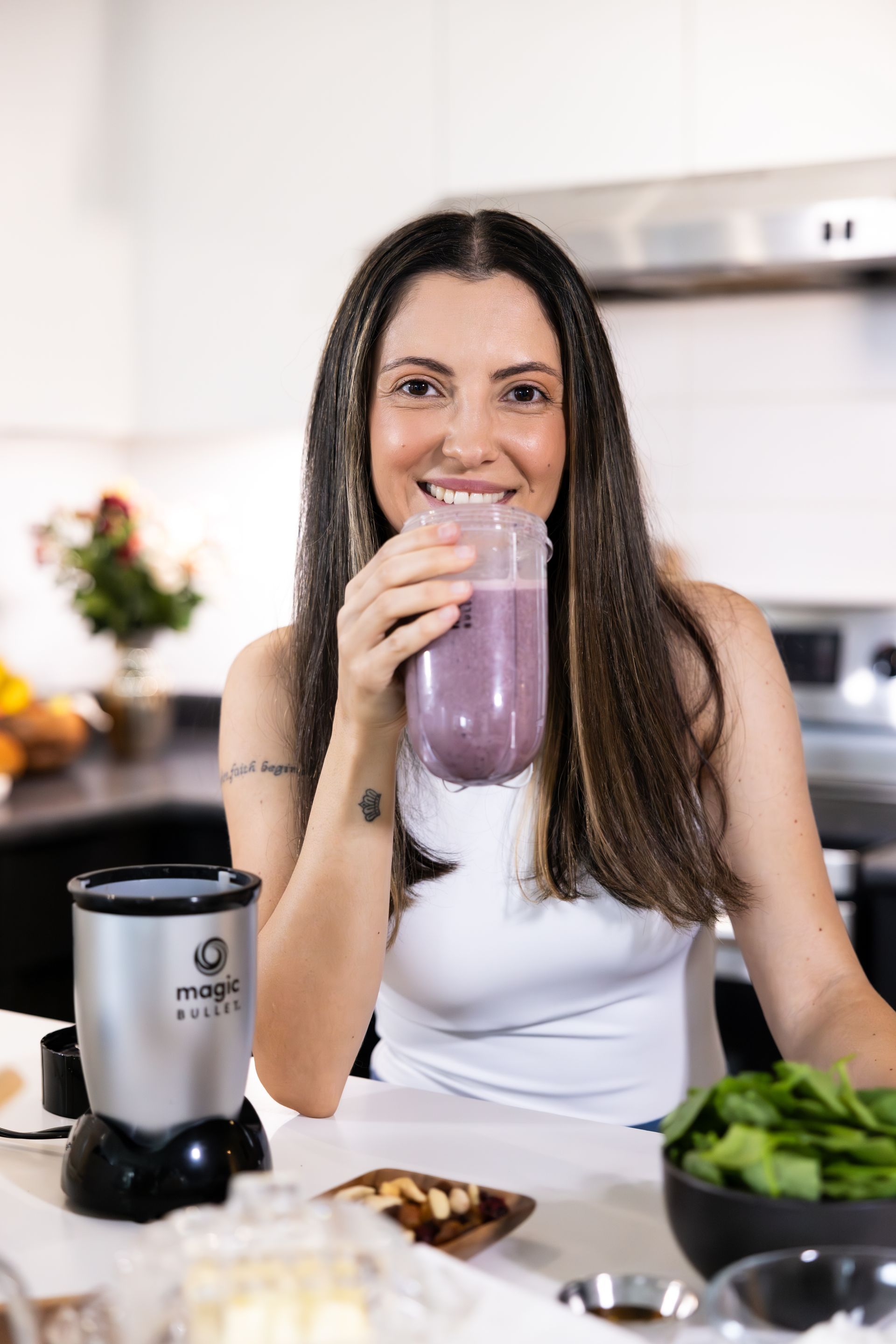 Woman in a kitchen drinking a purple smoothie beside a blender and fresh greens