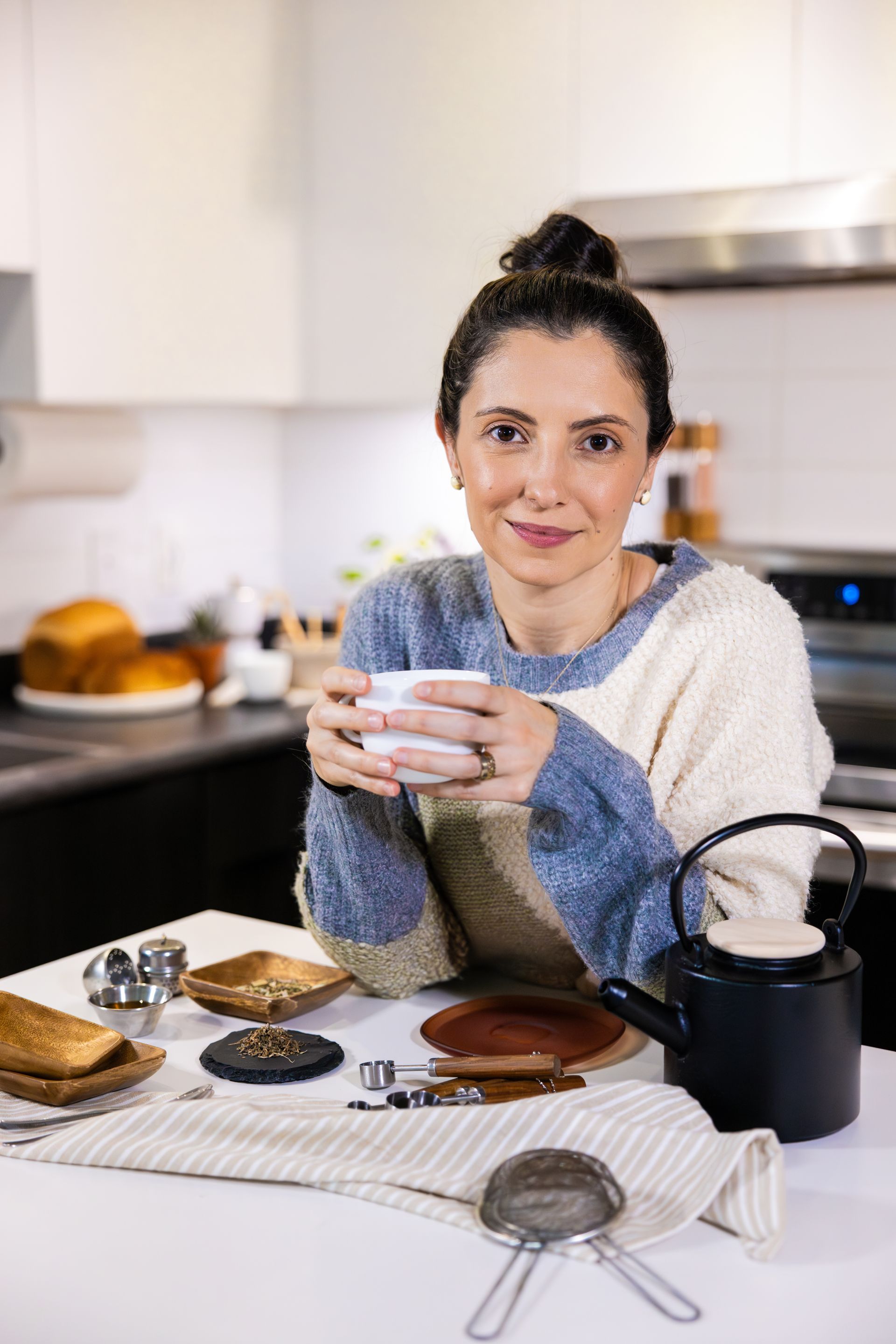 Woman holding a mug at a kitchen counter with pastries and a teapot.