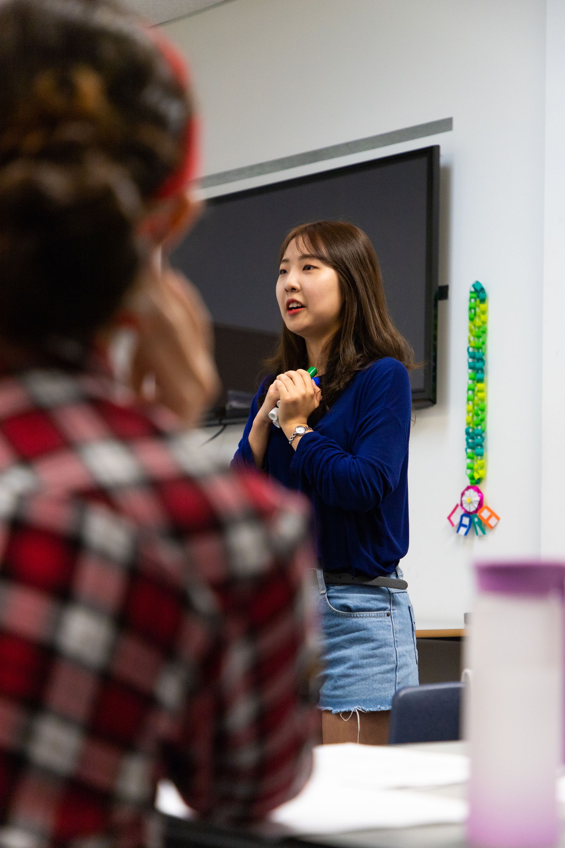Woman speaking in a classroom, wearing a blue sweater and denim shorts, with a blurred audience in the foreground