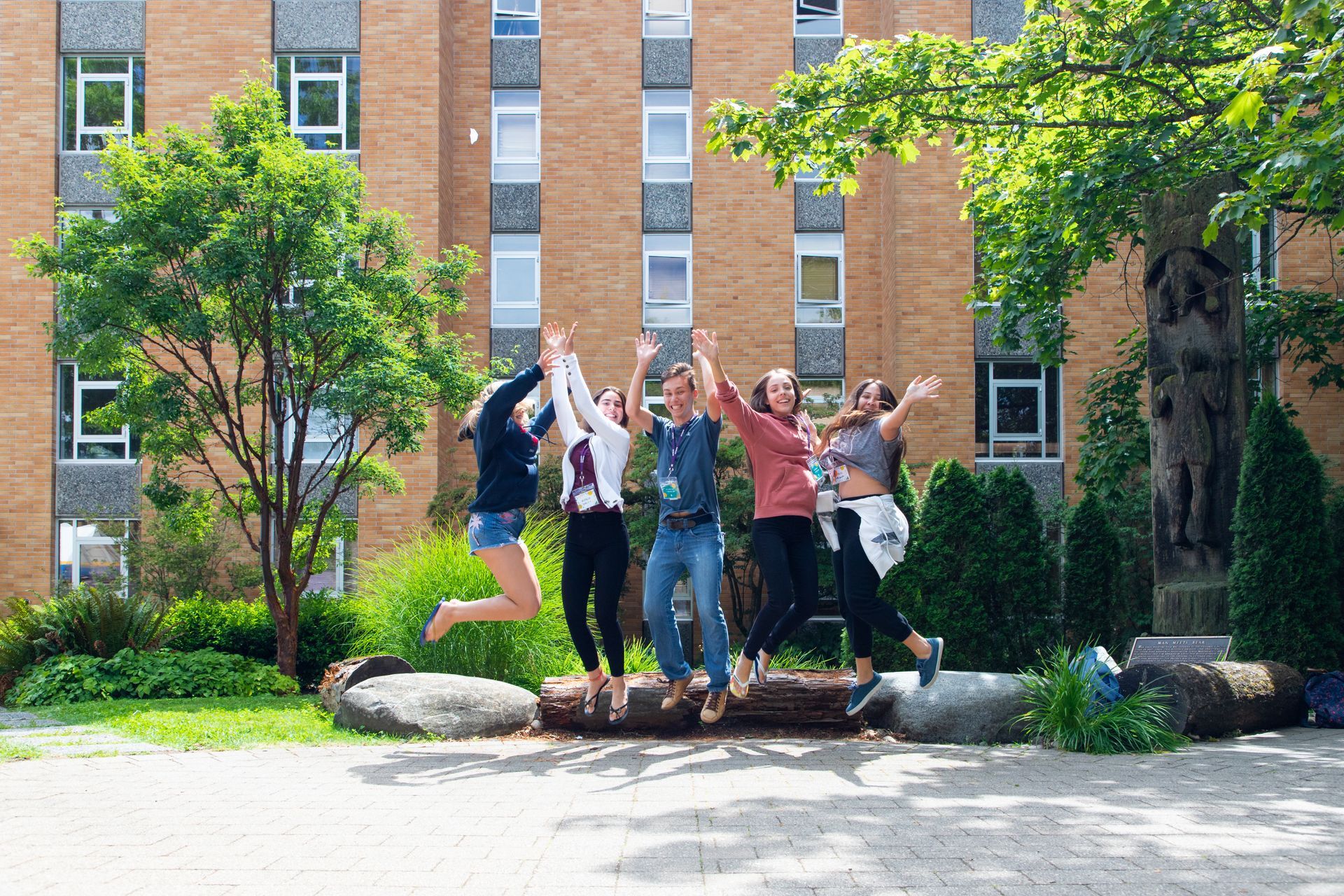 Group of people jumping together on a tree trunk in front of a brick campus building