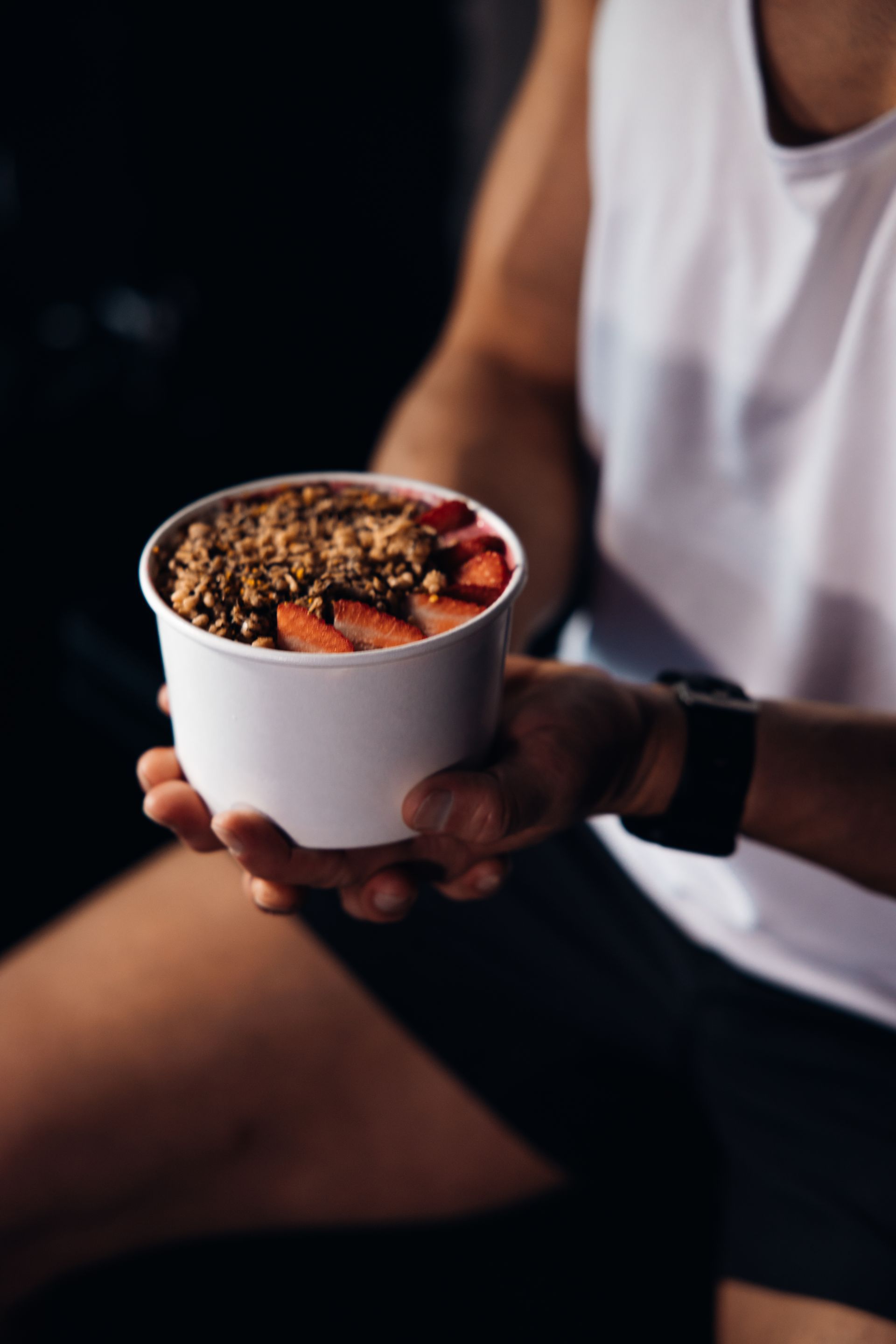 Person holding a white bowl of granola with strawberries in a dark workout setting