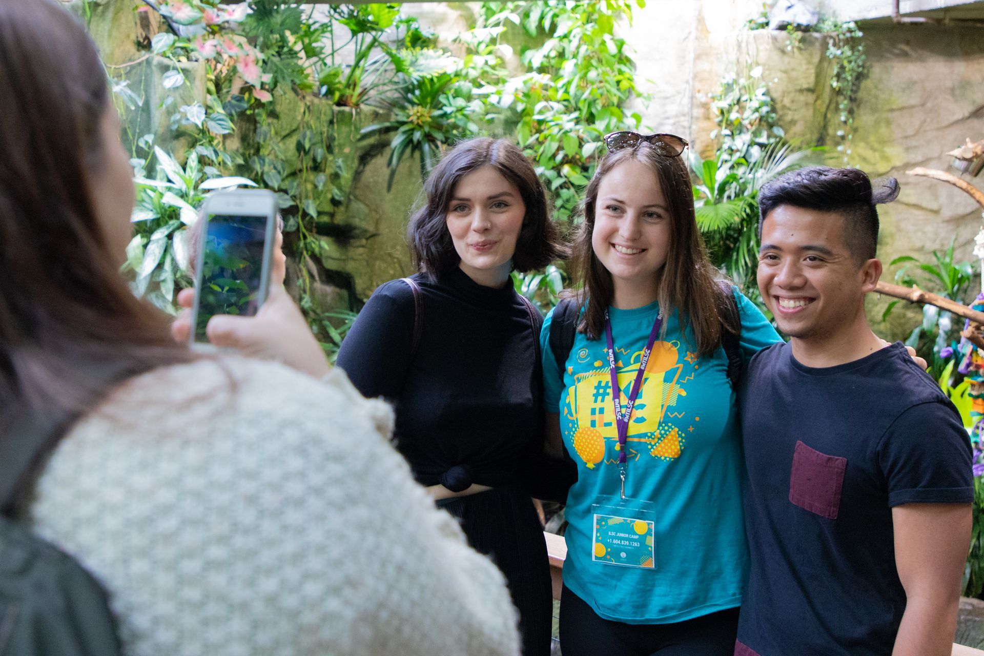 Three people pose outdoors while a person in the foreground takes their photo with a phone.