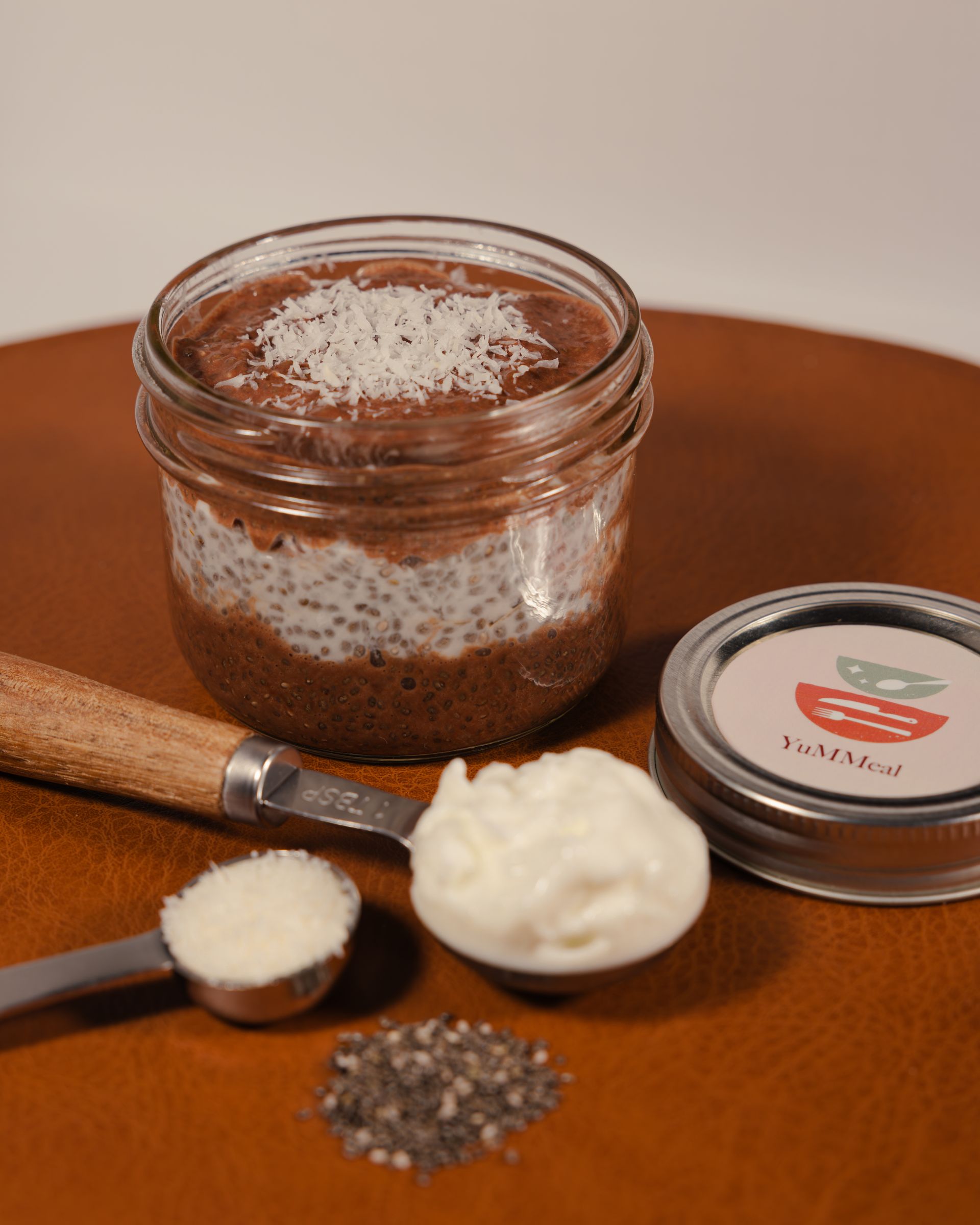 Open jar of brown scrub with spoon, cream dollop, and lid on a wooden table