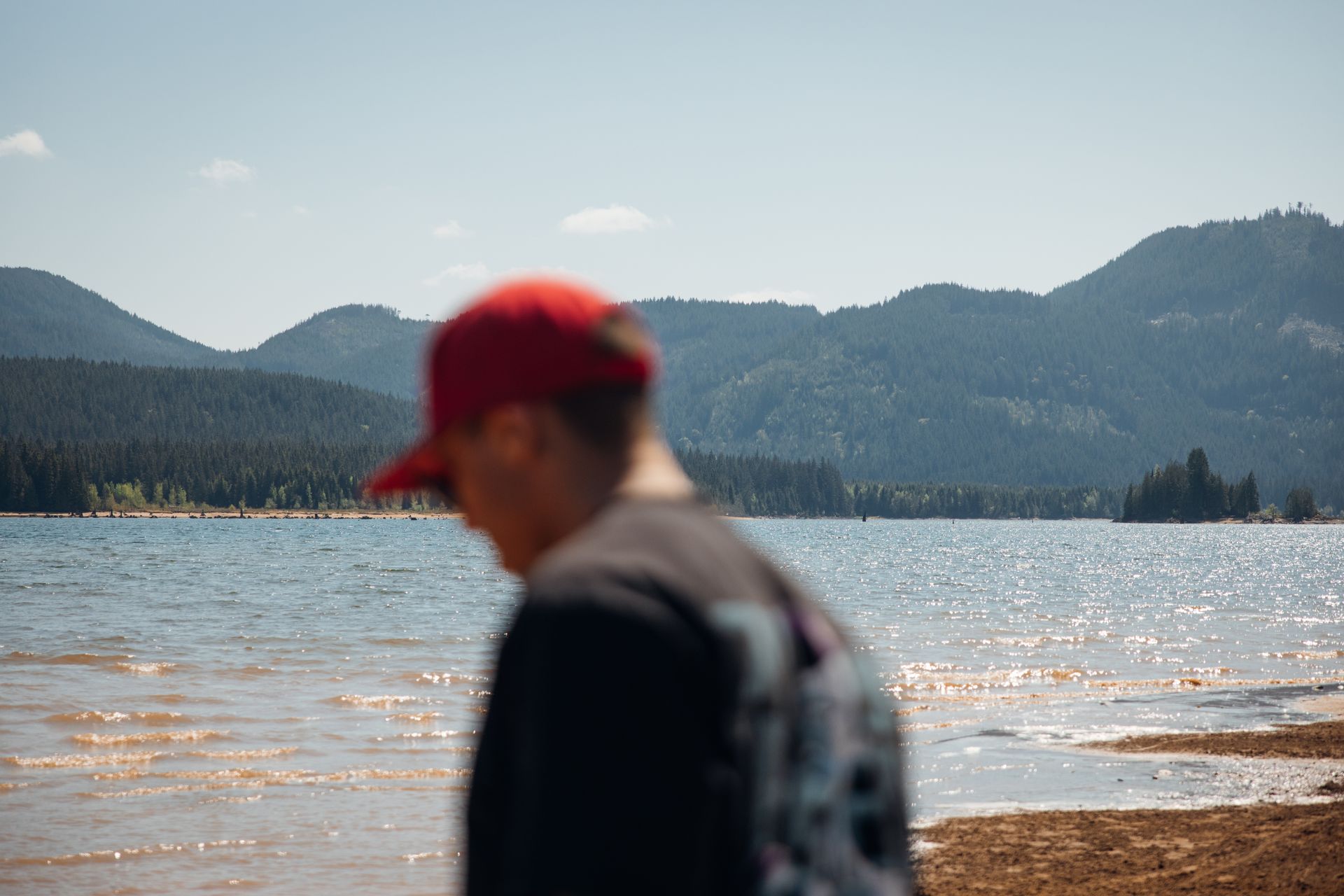 Blurred person in a red cap by a sparkling lake with forested mountains in the background