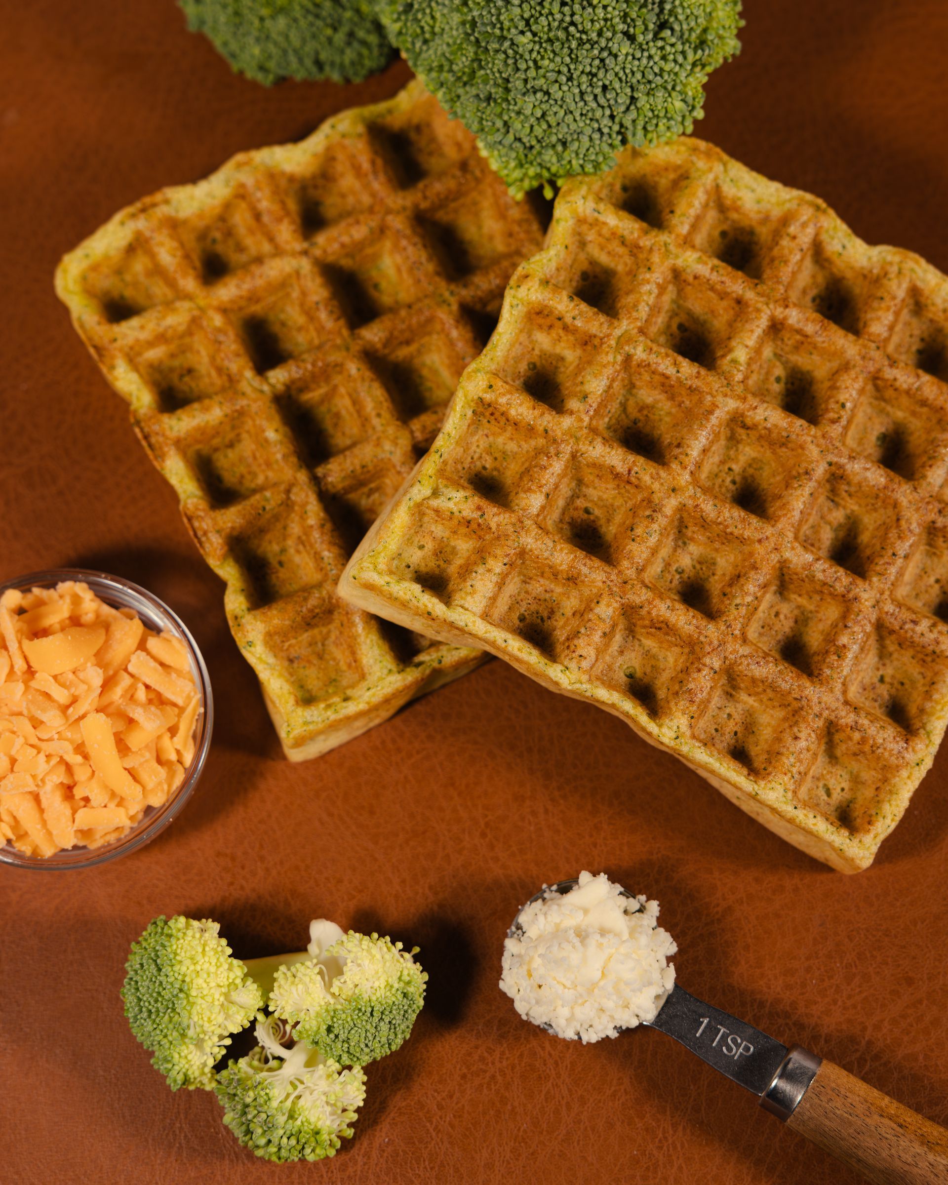 Two golden waffles with broccoli and grated cheese on a brown surface, with a spoon and a bowl of cheese.