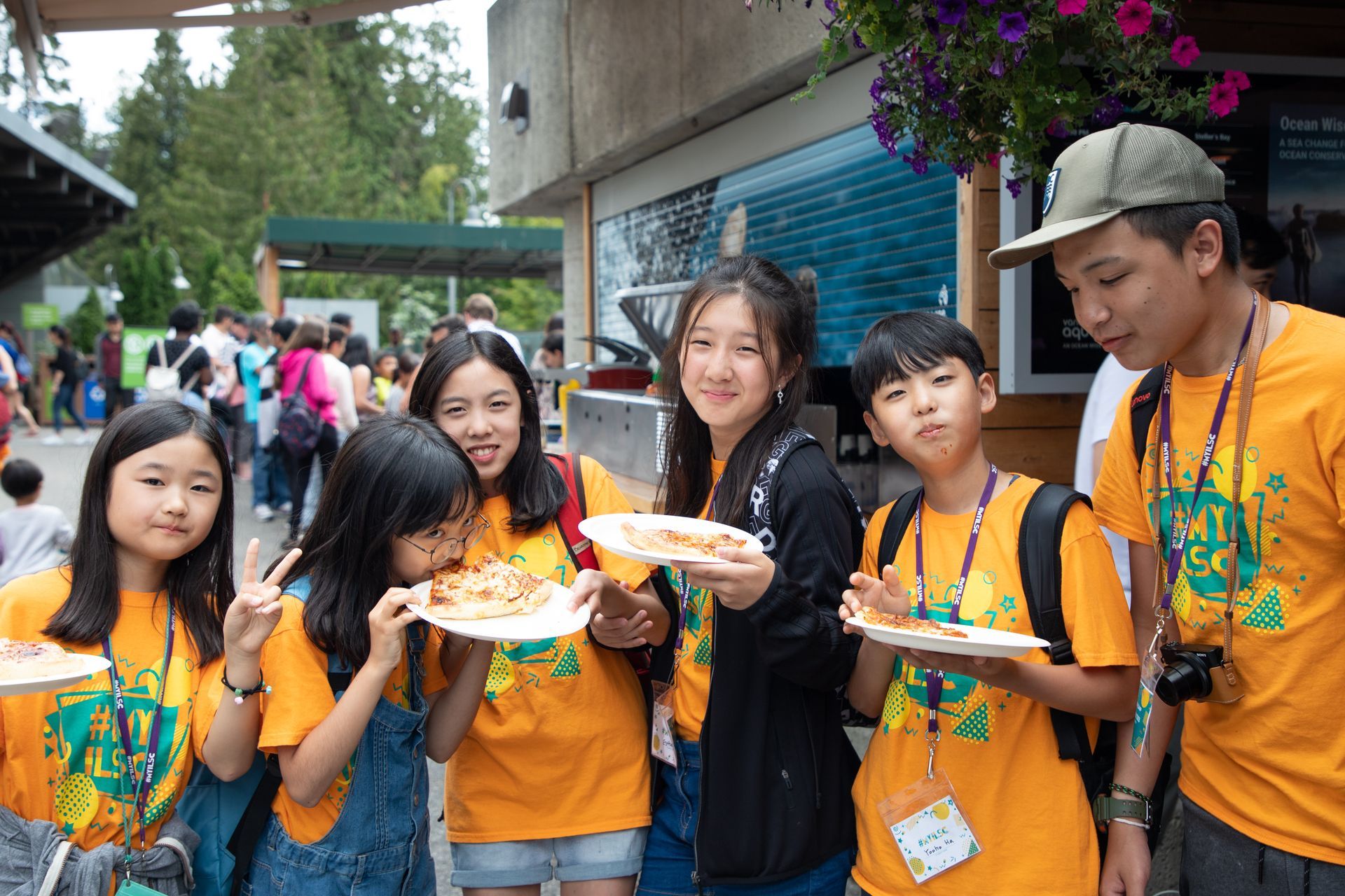 Group of children in orange shirts holding plates of food at an outdoor event.