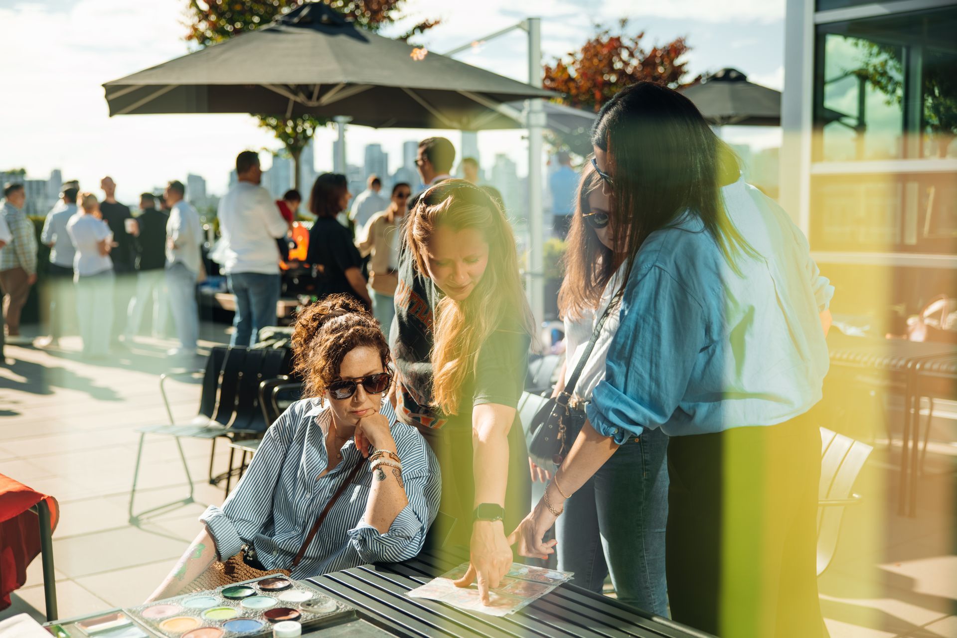 People gathered on a sunny rooftop patio, chatting around tables and a large umbrella.