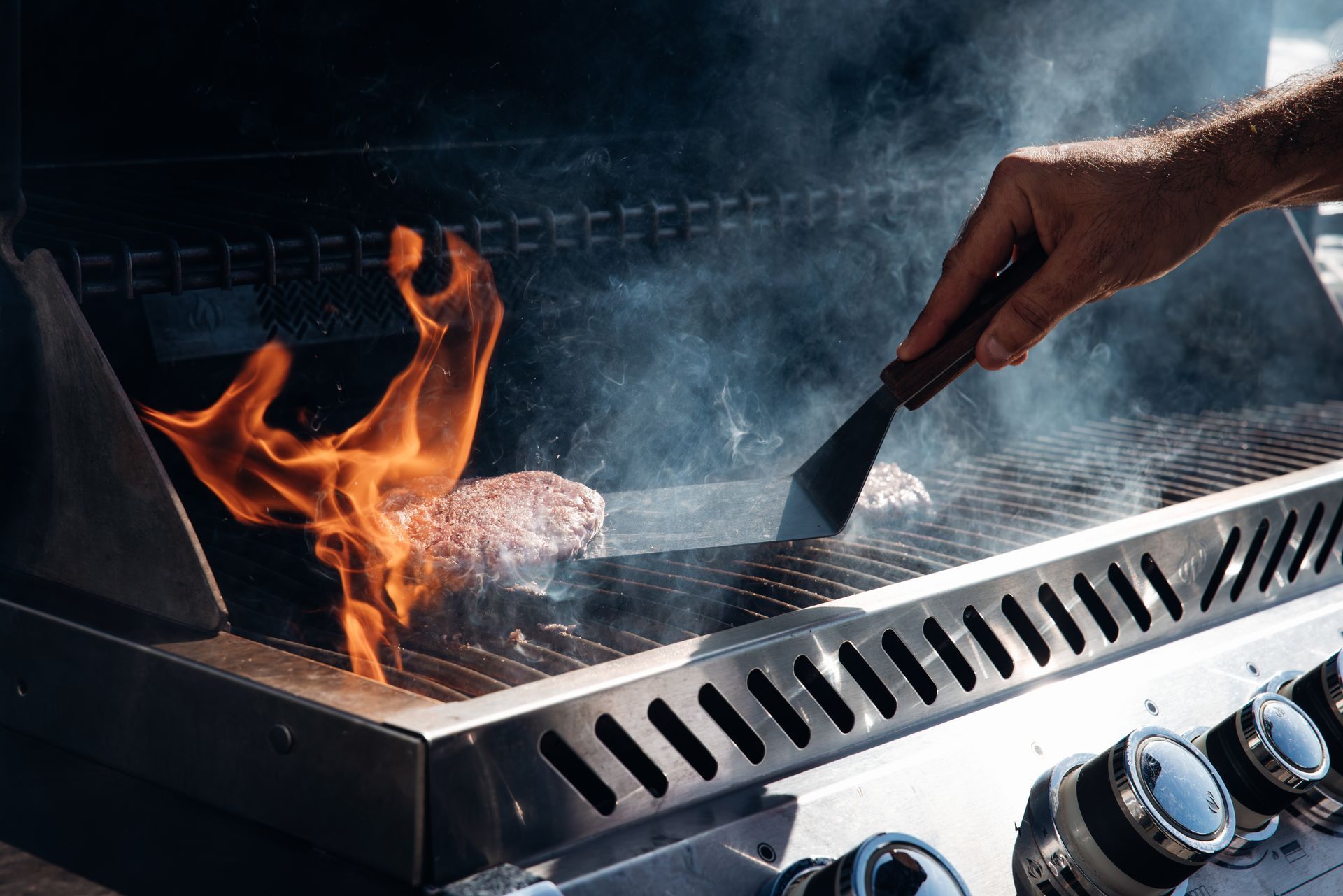 Hand stirring food on a flaming grill with smoke rising and control knobs visible