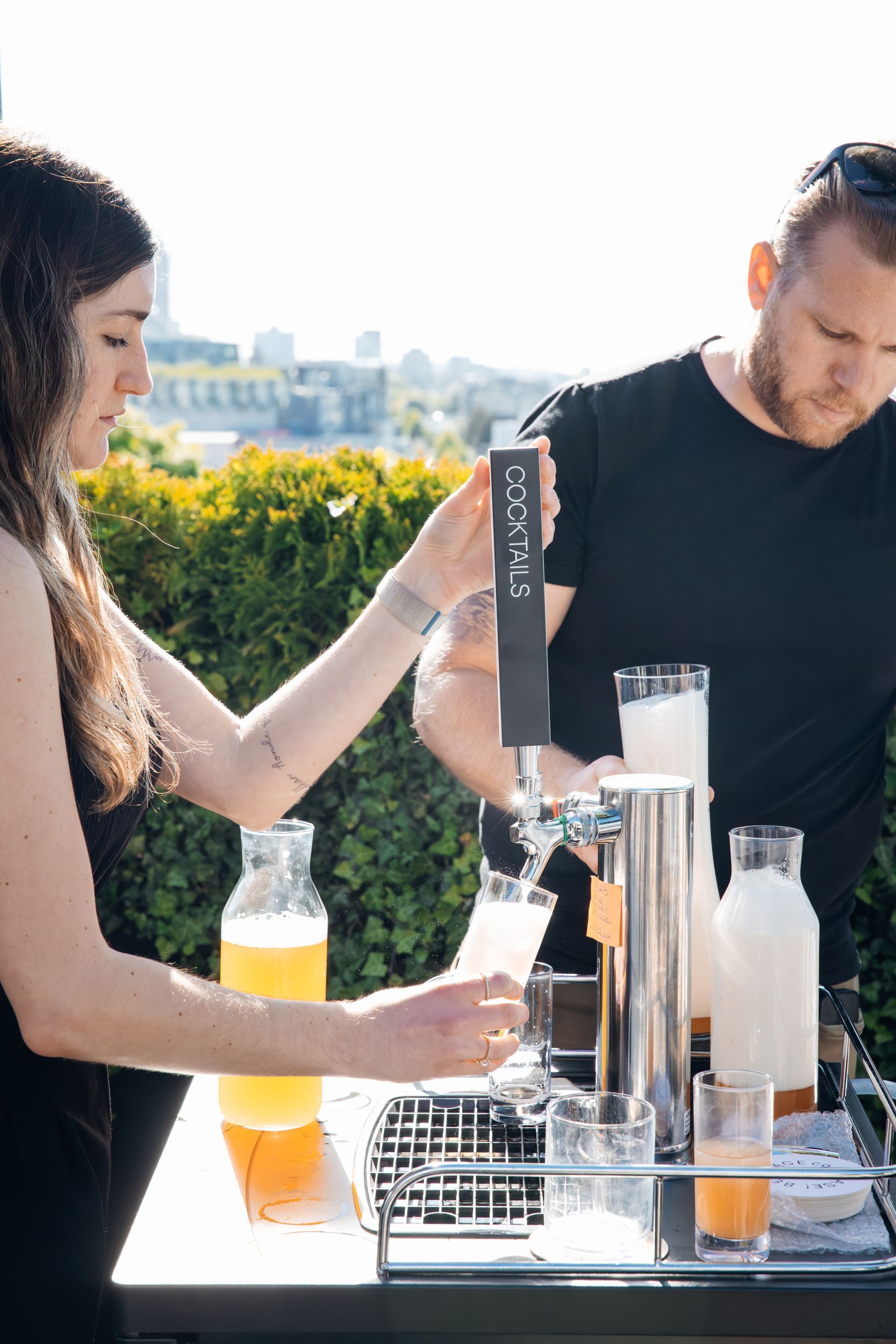 Two people preparing drinks at an outdoor bar with bottles, glasses, and a cocktail dispenser.