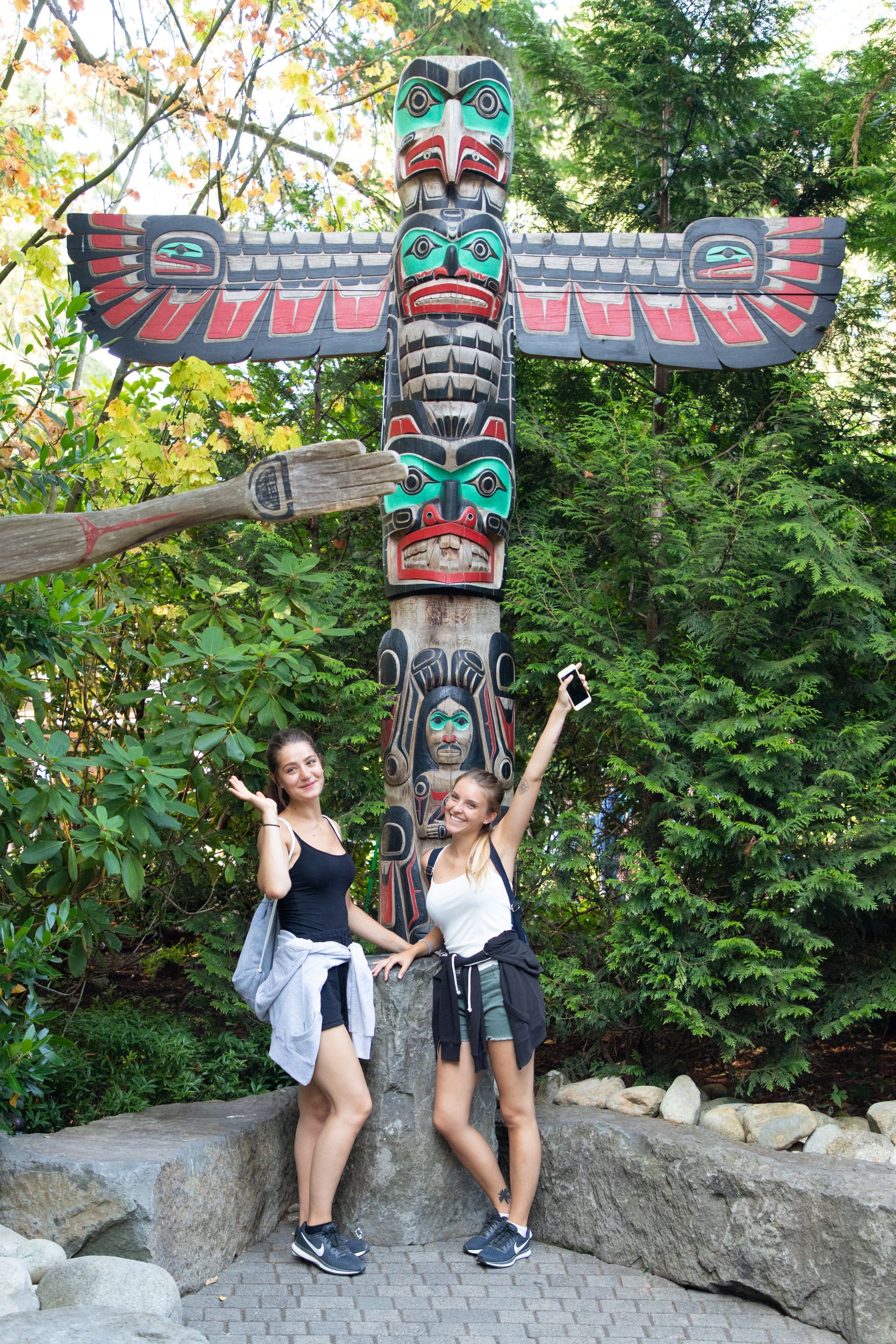 Two people posing in front of a colorful carved totem pole in a wooded park