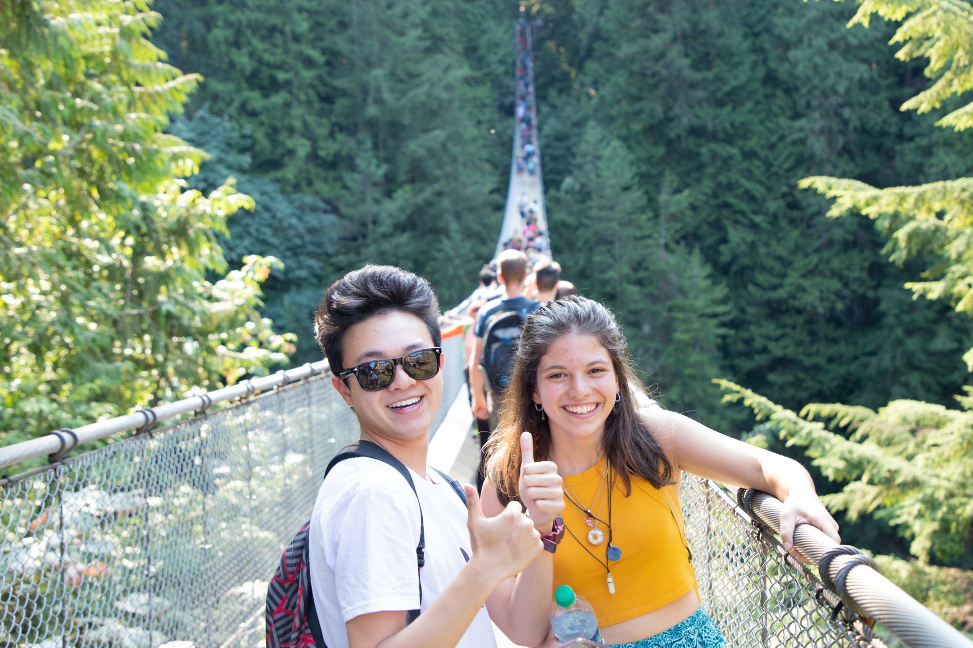 Two friends smiling on a suspension bridge in a forest, one giving a thumbs-up.