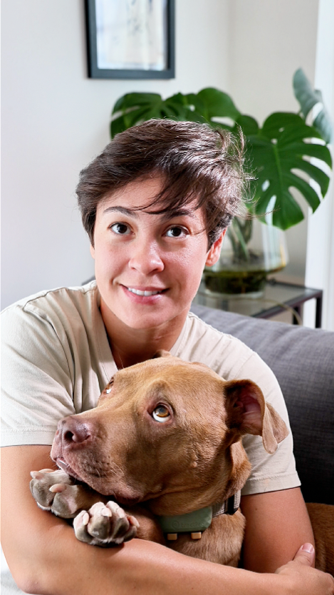 Person hugging a brown dog on a couch in a bright room with a large green plant in the background