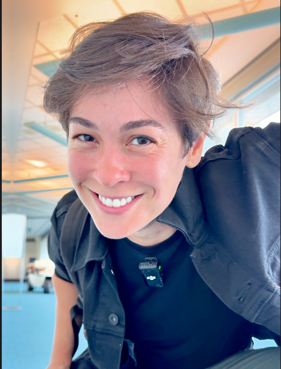 Smiling person leaning toward the camera in a bright indoor space, wearing a black shirt.