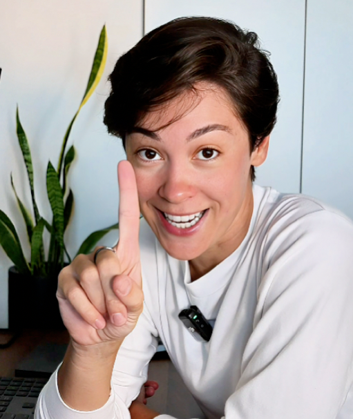 Person in white shirt smiling and holding up one finger indoors, with a plant in the background