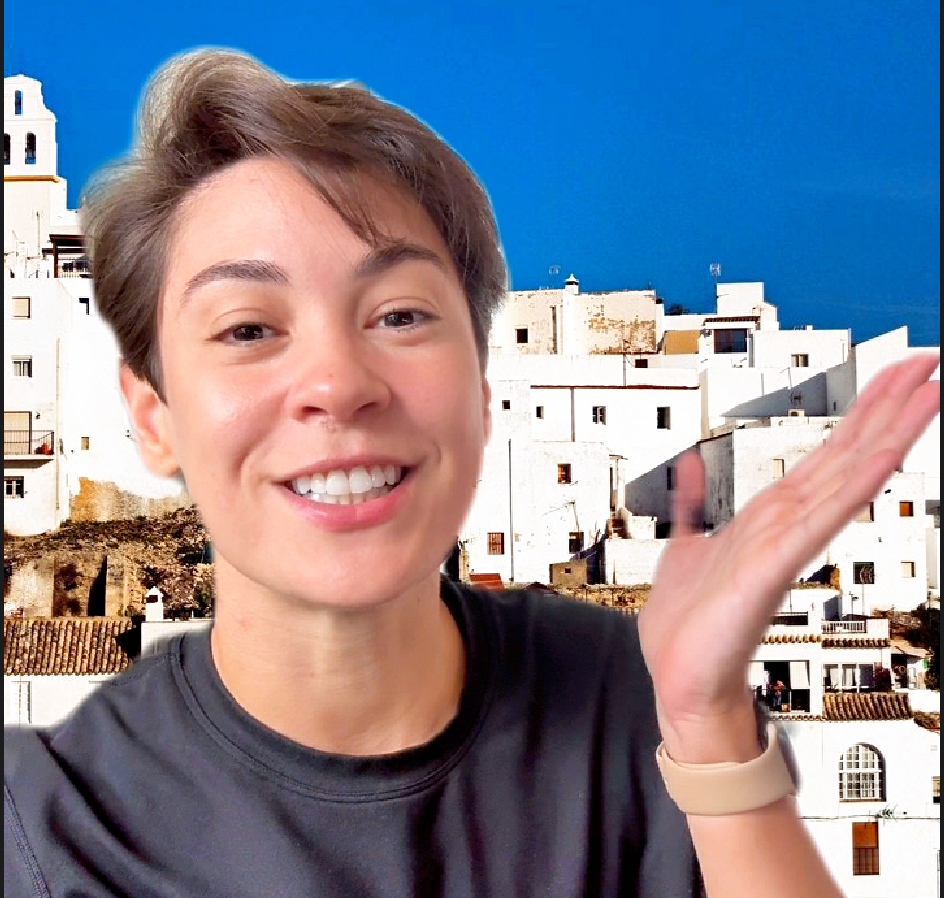 Smiling person waving in front of white buildings and a bright blue sky