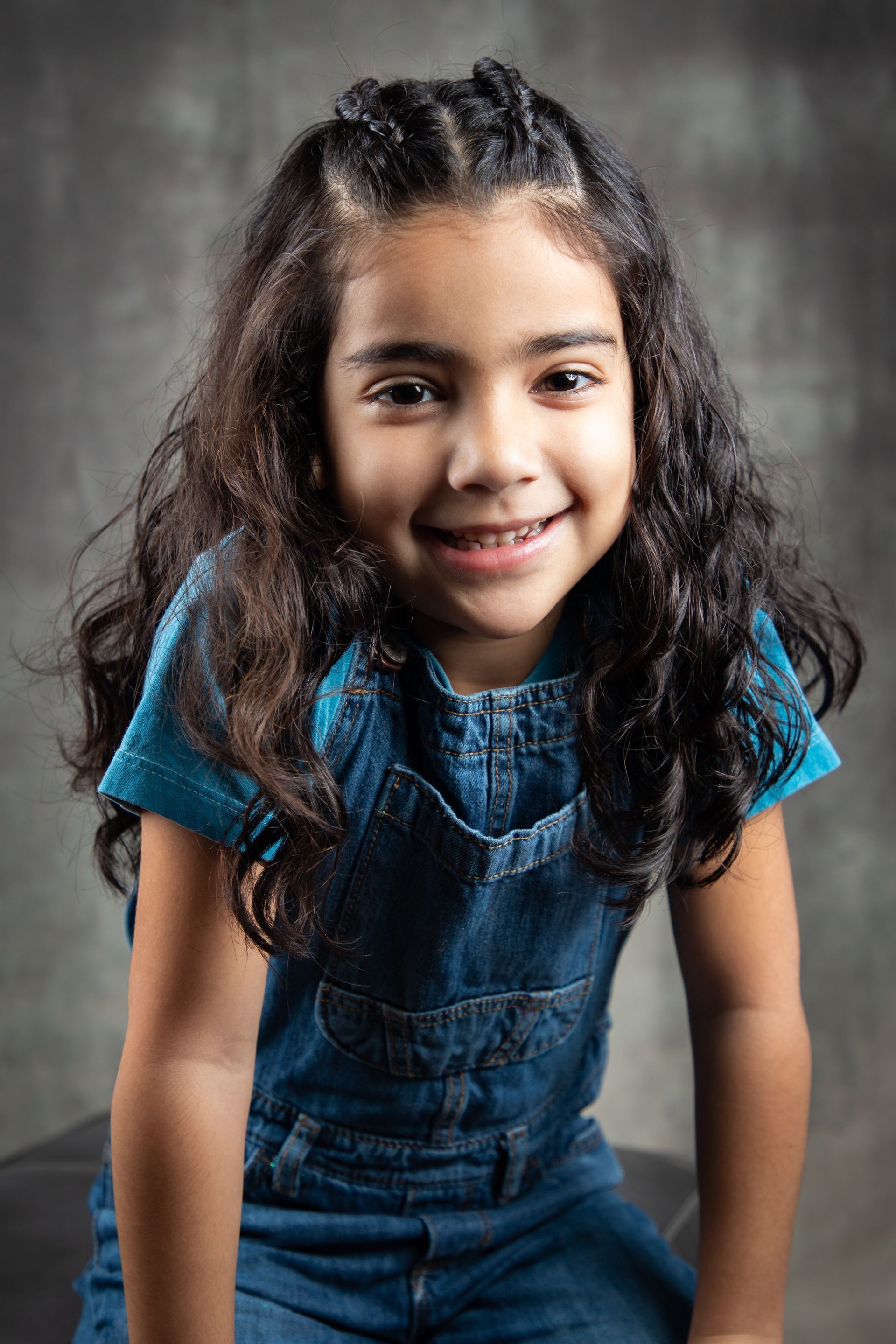 Smiling child in blue denim outfit, leaning forward against a gray studio backdrop