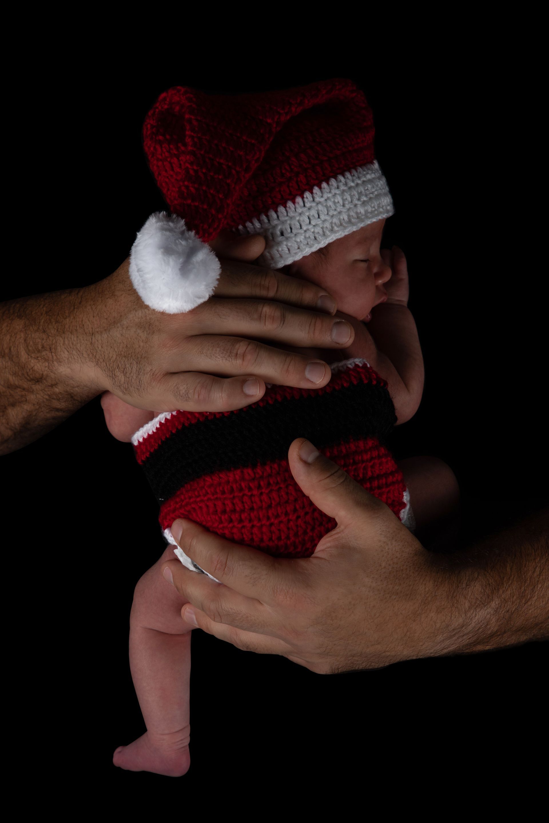 Baby in red knit outfit and hat, held gently by adult hands against a black background
