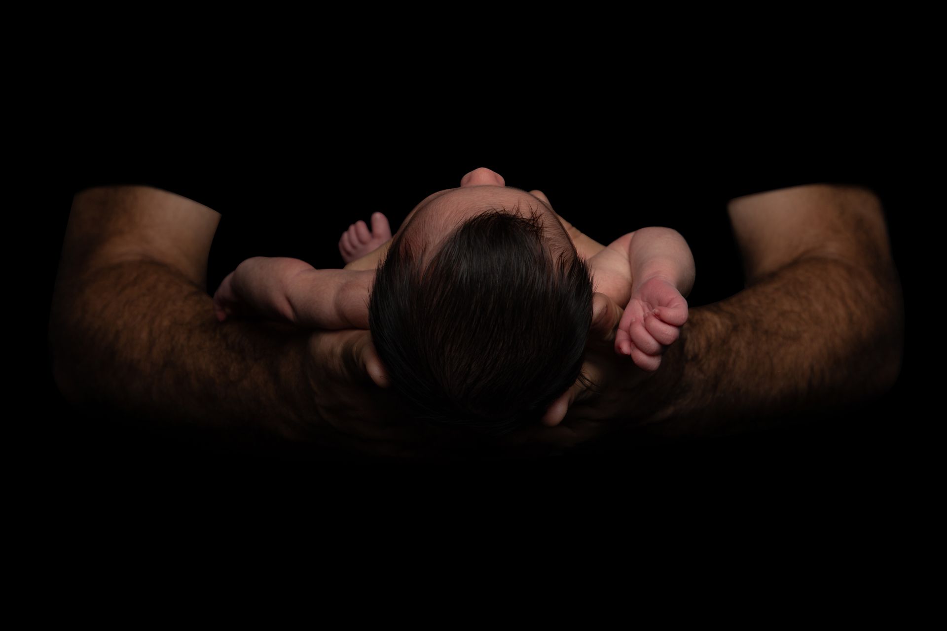 A newborn lying on an adult’s arms against a dark background, hands near its face.