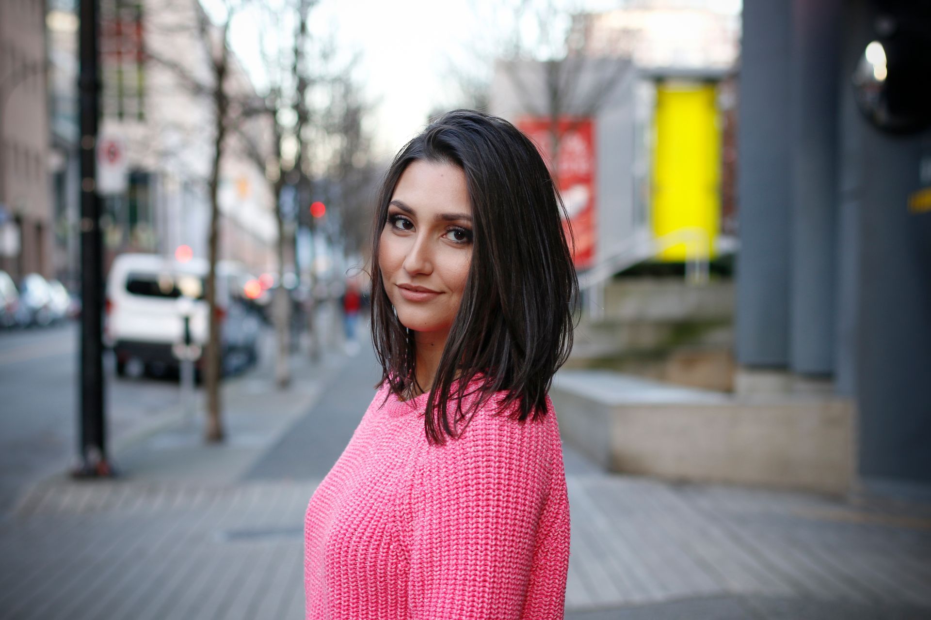 Woman in a bright pink sweater standing on a city sidewalk, looking at the camera.