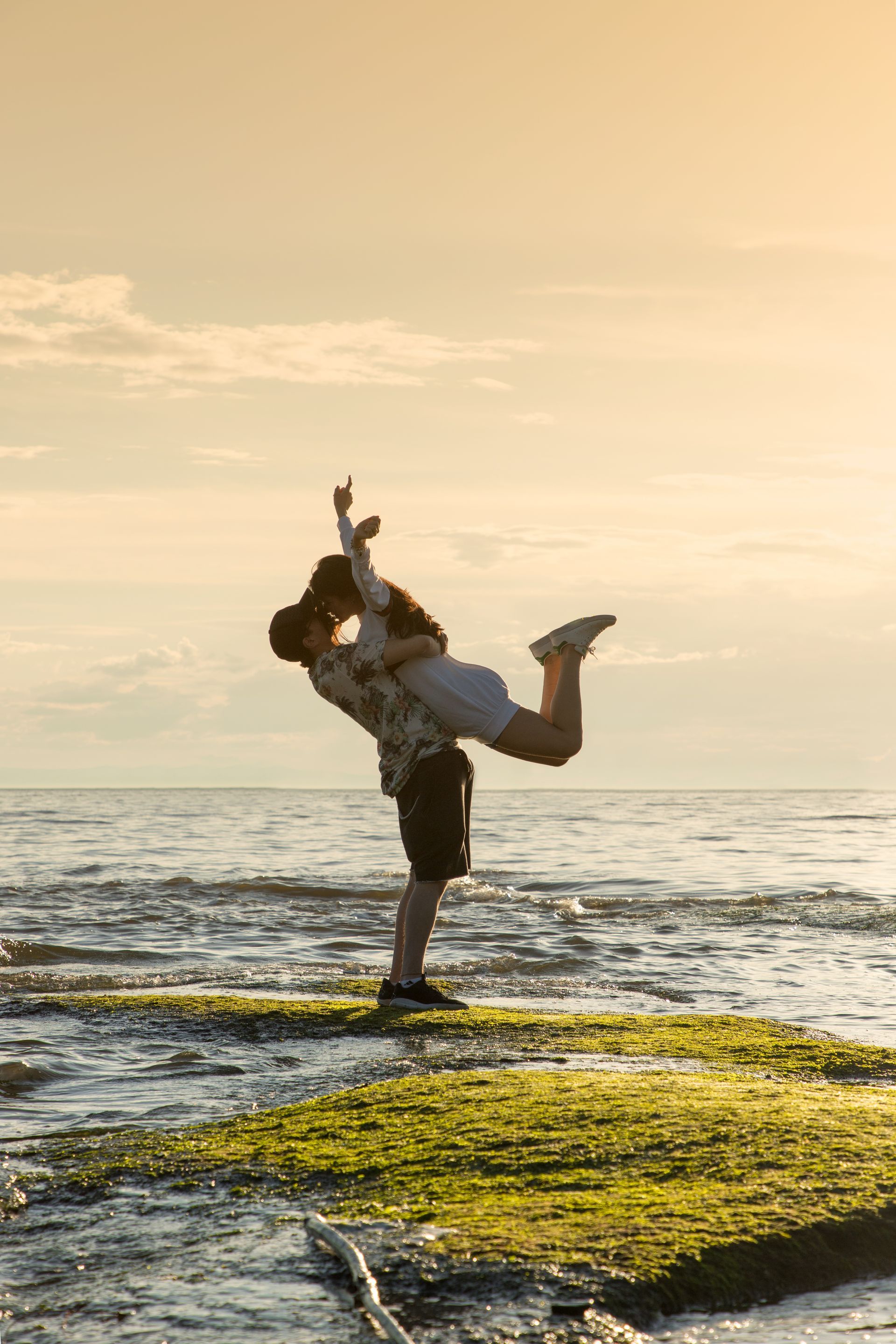 Person lifting another person on a rocky shore at sunset with ocean in the background