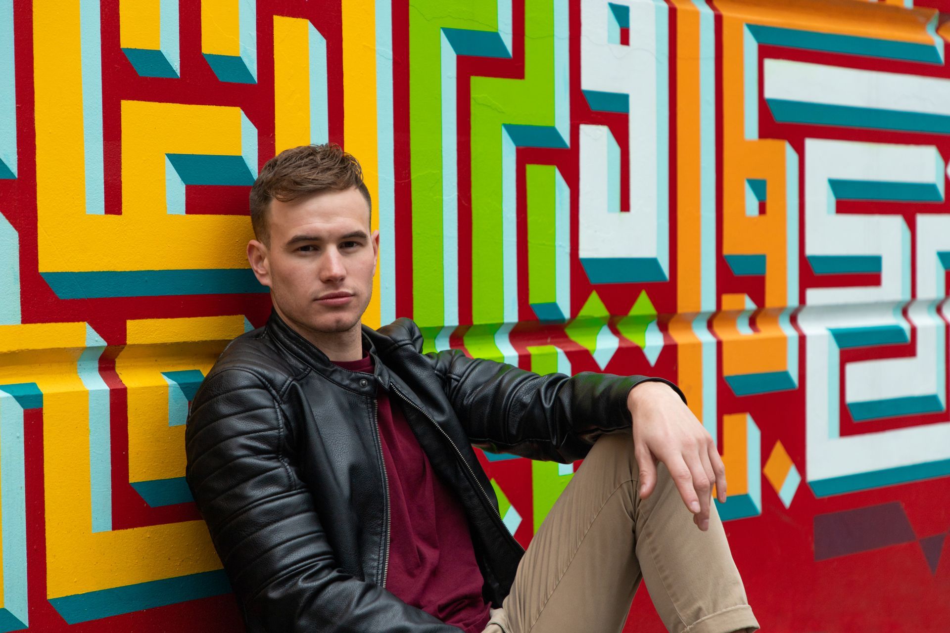 Man sitting by a colorful geometric mural, wearing a black jacket and tan pants
