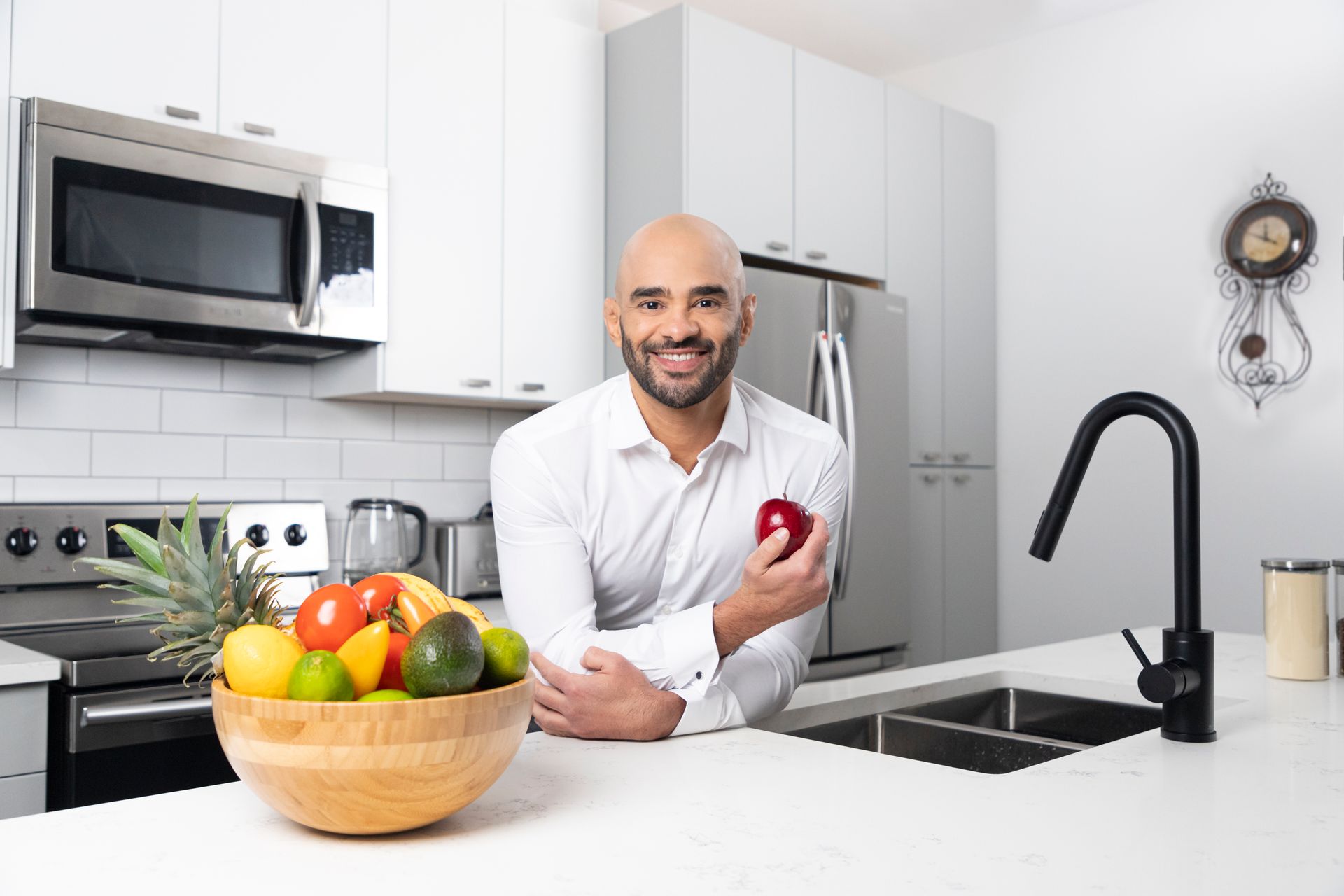 Smiling person in a white kitchen with a fruit bowl, stove, microwave, and black faucet
