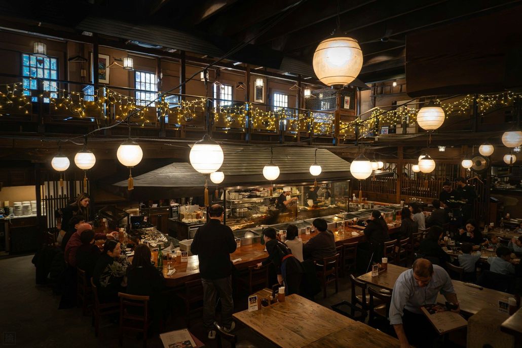 Busy warmly lit restaurant interior with hanging lanterns, crowded tables, and diners eating and talking.