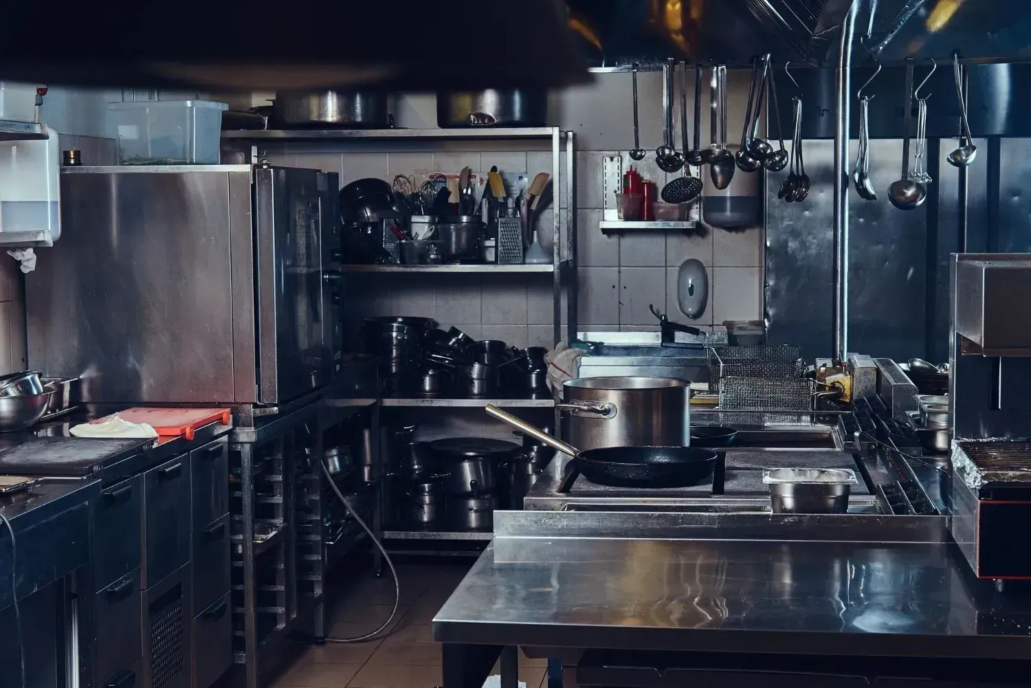 Stainless steel commercial kitchen with pots, pans, and utensils hanging above a worktable.